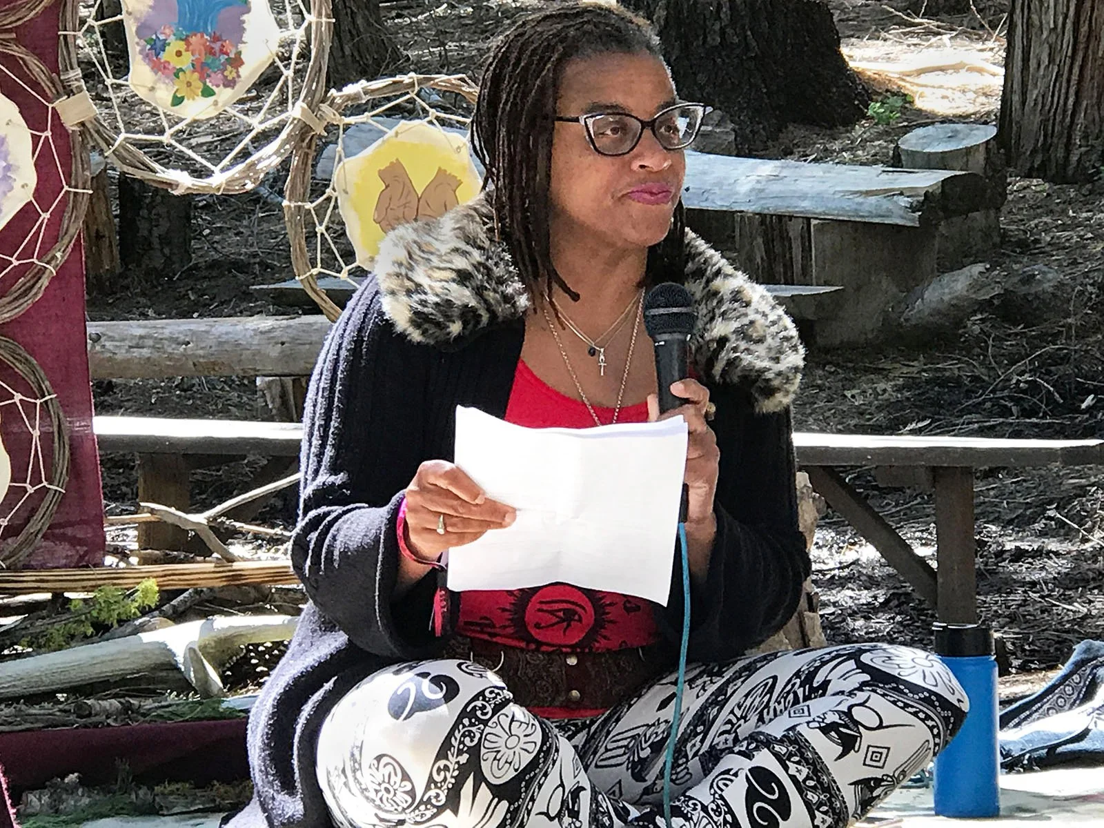 Ali Montgomery speaks to a gathered community in an outdoor woodland setting, holding a microphone and notes. Behind her, sybolic artworks form a backdrop, evoking themes of ritual, embodiment, and cultural storytelling.