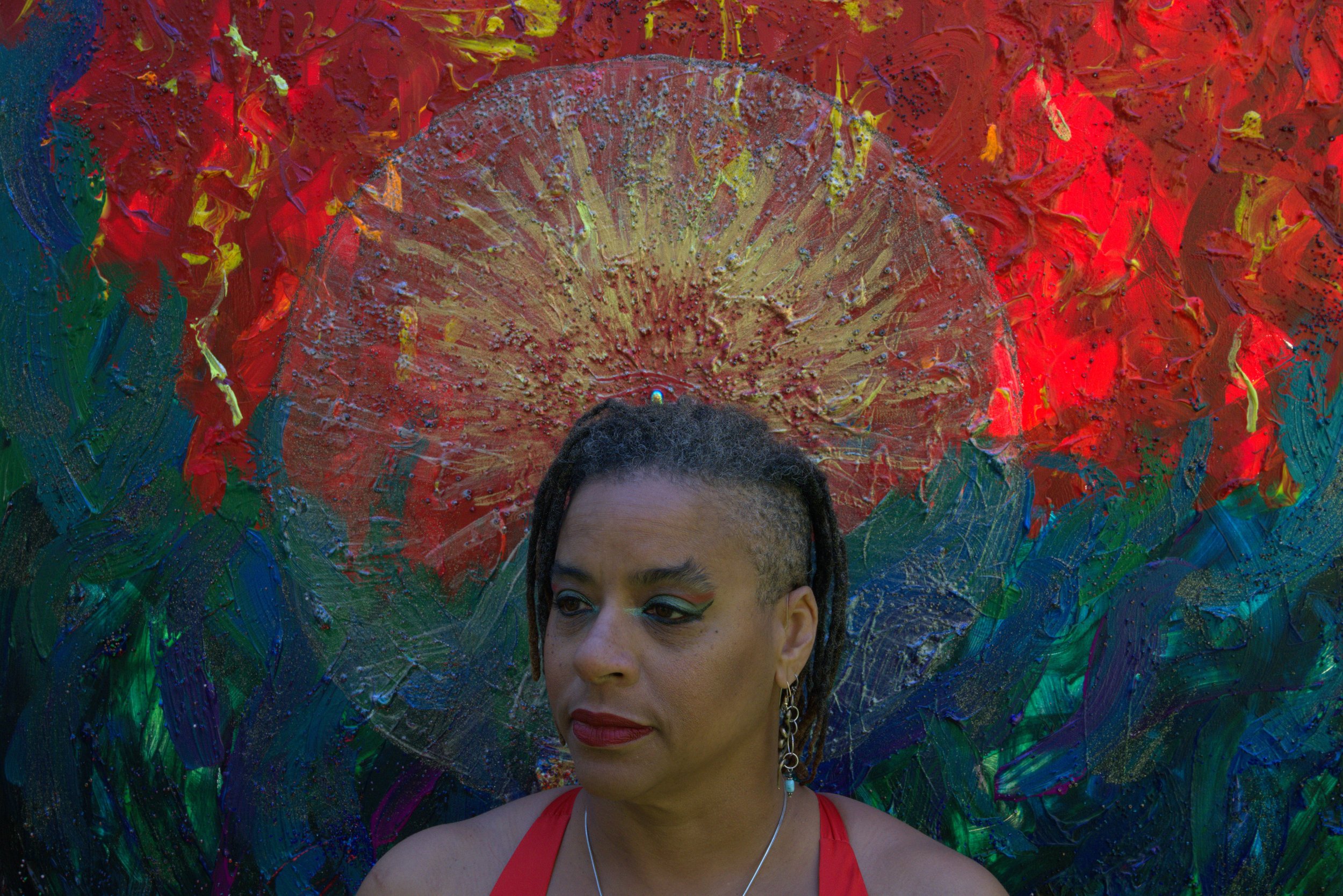 Portrait of Ali Montgomery standing before her painting “Ascending the Priestess Path,” with radiant circular strokes in red, yellow, green, and blue. Her red halter top and bold makeup echo the artwork’s vibrancy, creating a visual fusion of prieste