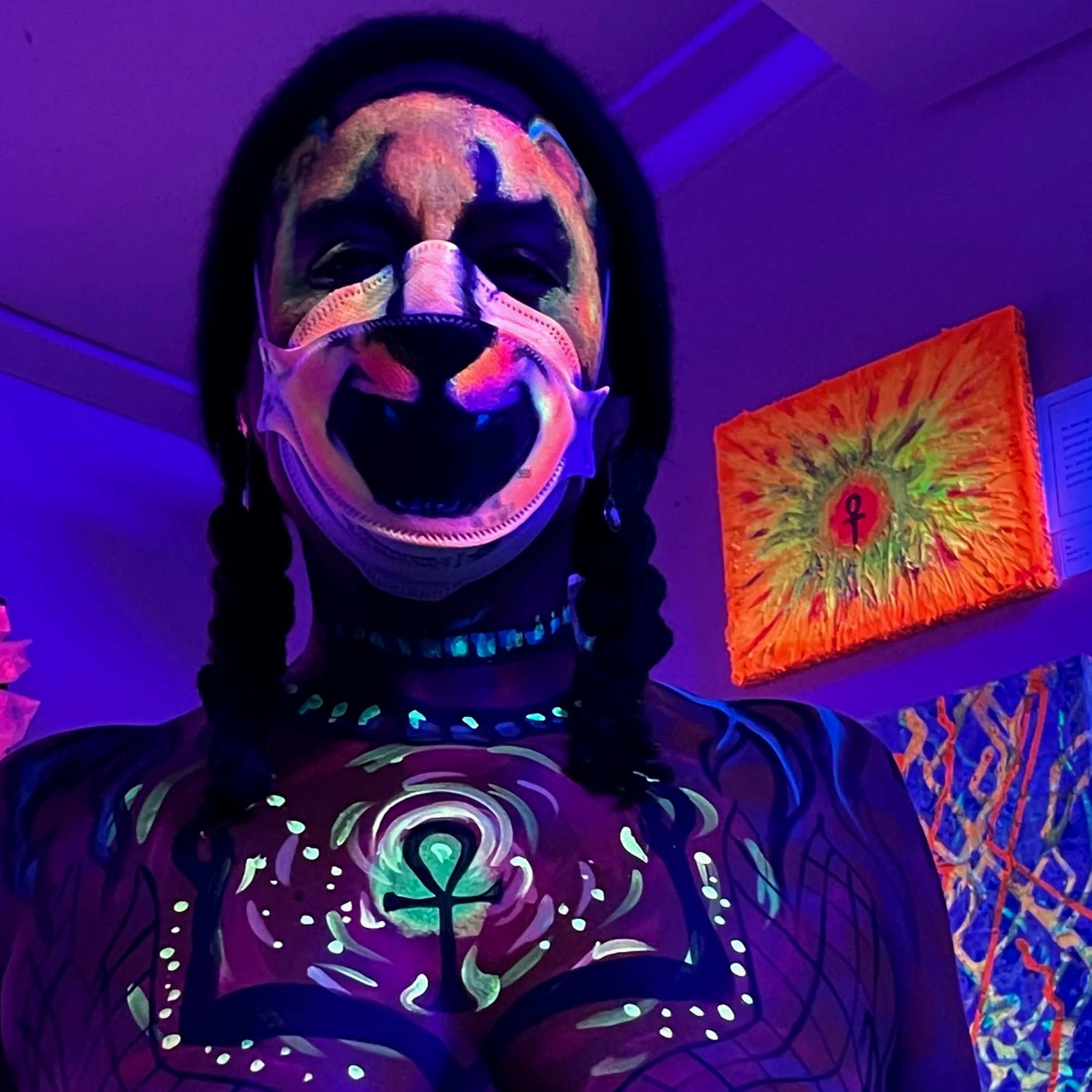 Ali Montgomery dancing before her painting “Sol’s Soul,” illuminated under black light. Her face is painted with lioness features, and her chest is painted with a glowing ankh.