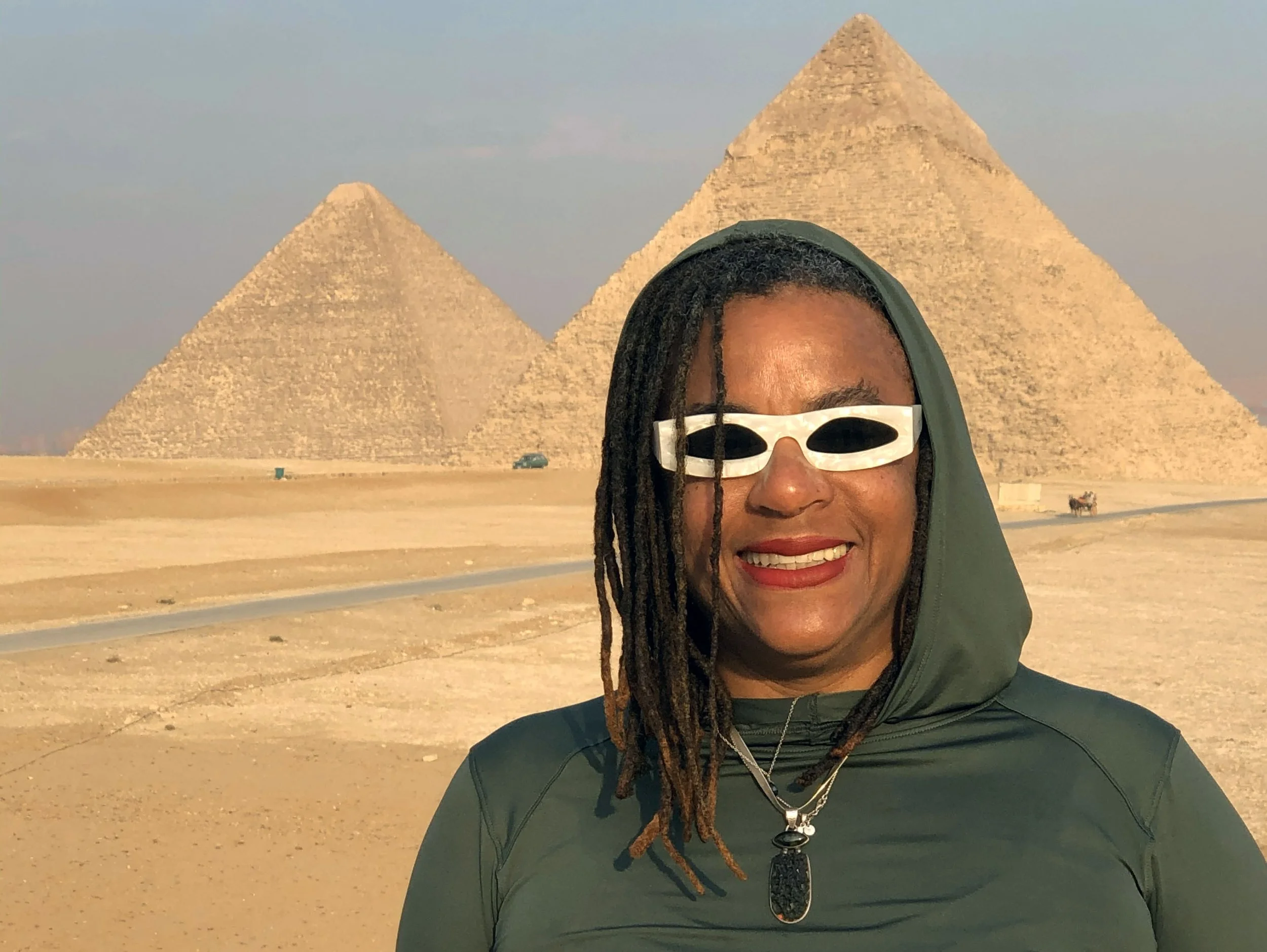 Ali Montgomery wearing white‑framed sunglasses and dreadlocks standing before the Great Pyramids at Giza, with a wide expanse of yellow desert between her and the pyramids.