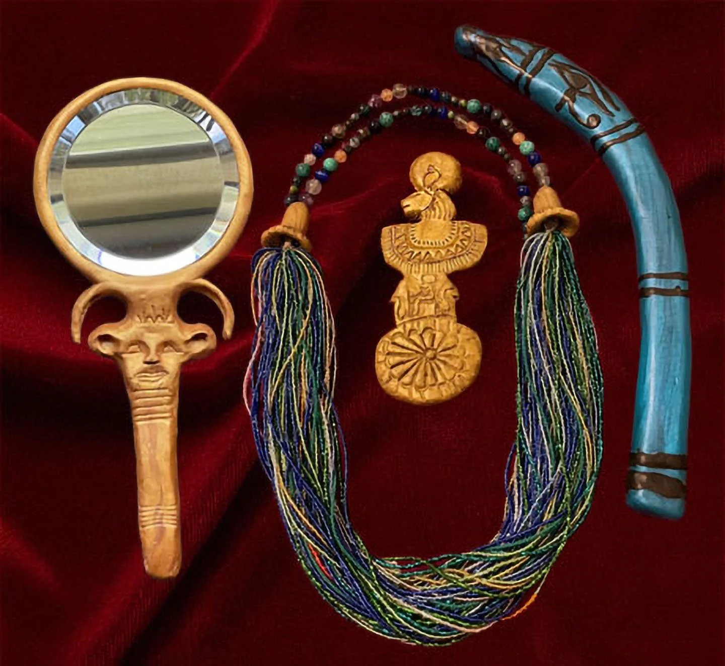 Collection of ritual-inspired objects arranged on red velvet fabric. Items include a multistrand menat necklace of blue, green, and purple beads with a gold pendant depicting the lion‑headed goddess Sekhmet, a gold‑handled mirror with a Hathor‑head d