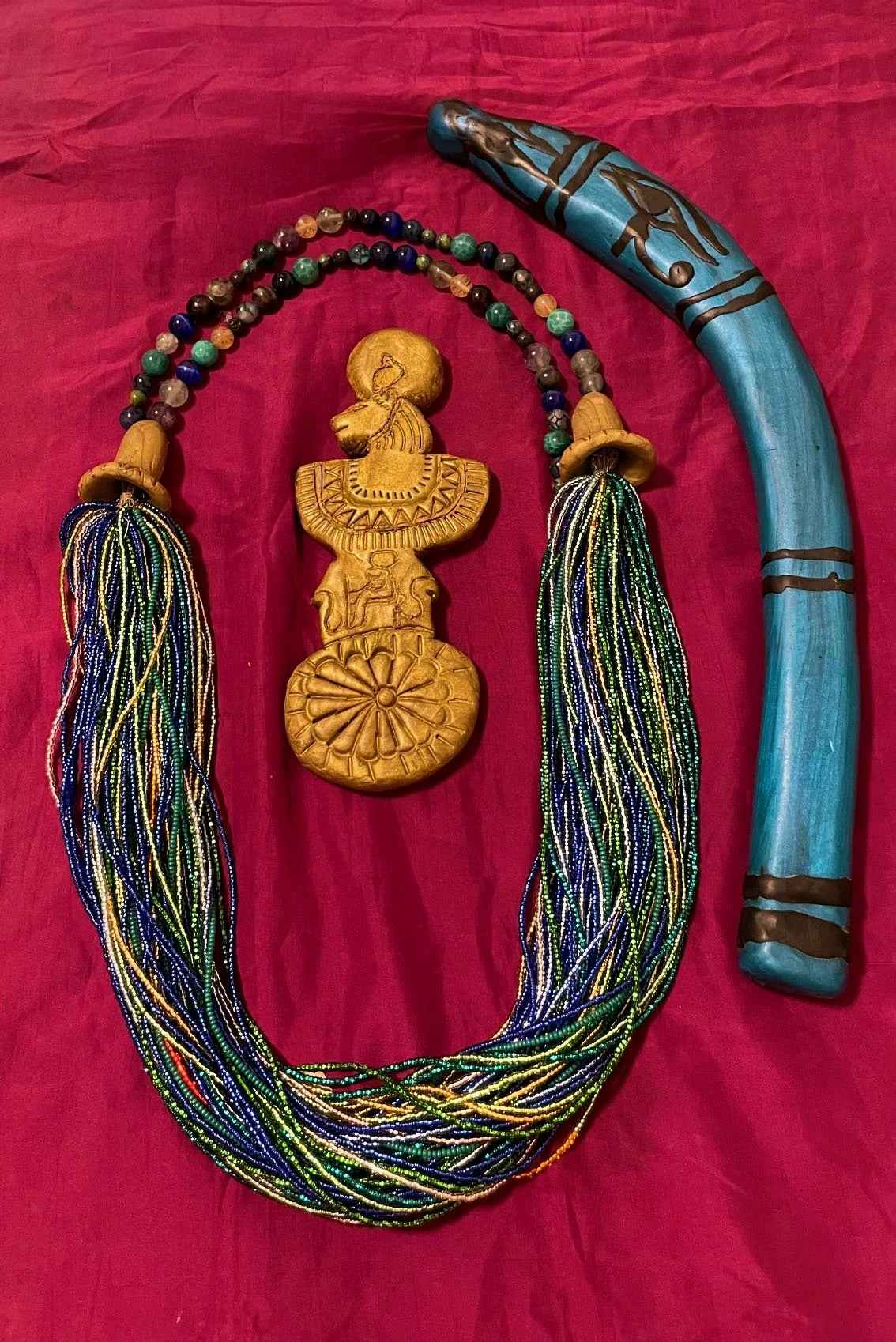 Ritual-inspired objects arranged on red fabric. Items include a multistrand menat necklace with small multicolored beads and larger spherical beads, featuring a golden pendant depicting the lion-headed goddess Sekhmet. Beside it lies a blue throw-sti