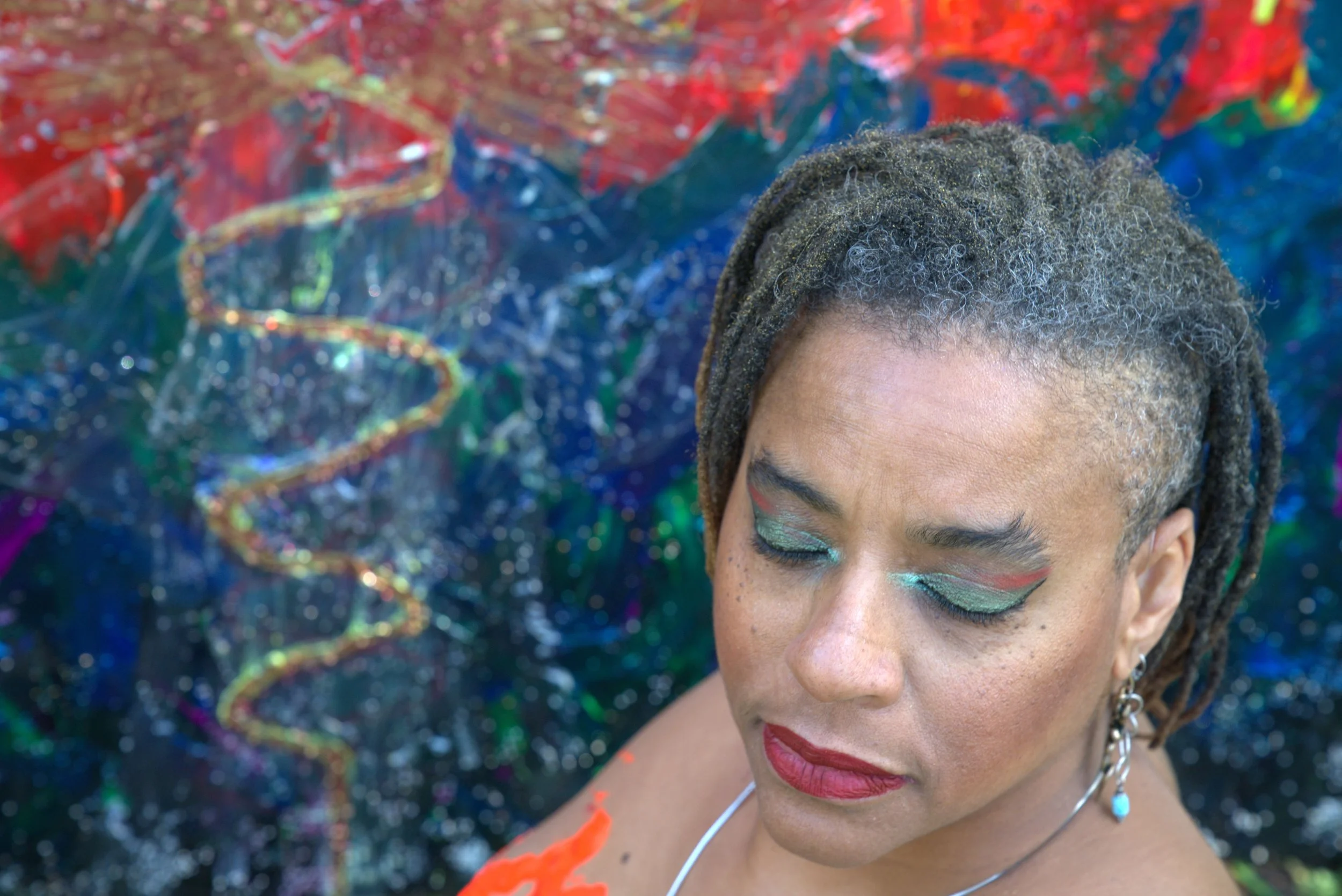 Ali Montgomery stands with closed eyes in front of her painting “Ascending the Priestess Path,” featuring a serpentine gold line rising across strokes of red, blue, and green. She wears bold makeup, short dreadlocks, and a necklace. 