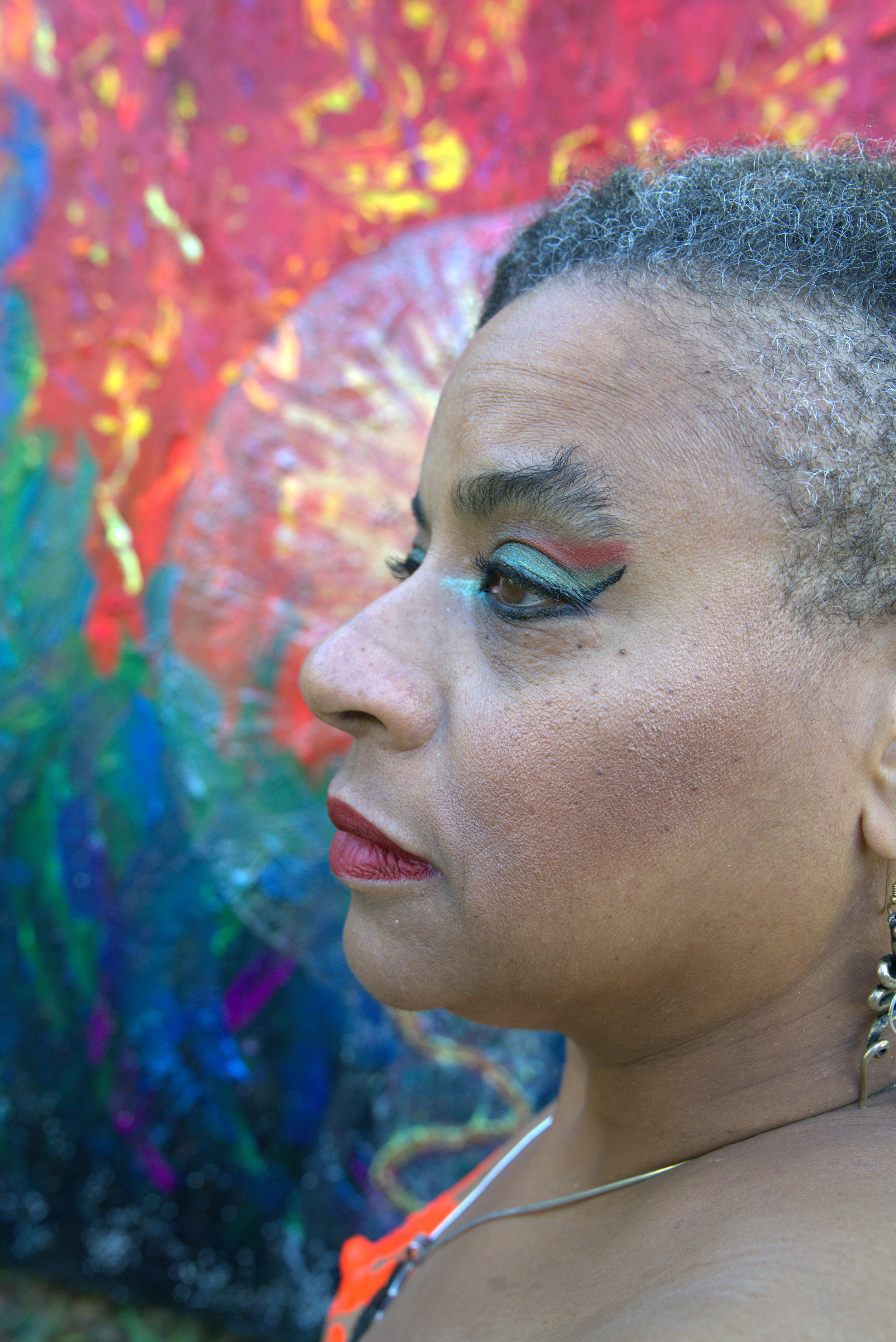 Close-up facial profile of Ali Montgomery with backdrop of her painting “Ascending the Priestess Path,” featuring swirling strokes of red, yellow, blue, green, and purple. She wears bold makeup and a necklace.
