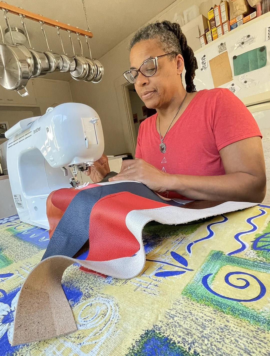 Ali Montgomery using a sewing machine to stitch leather pieces for a Nubian skirt replica in red, orange, blue, and white.