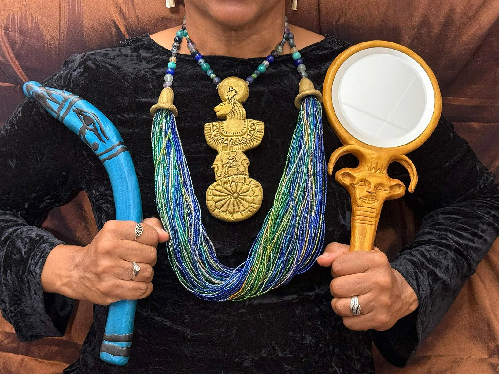 Menat necklace of blue and green seed beads with golden lioness pendant, blue curved stick with black udjat eye marking, and mirror with golden handle in shape of stylized Hathor face.