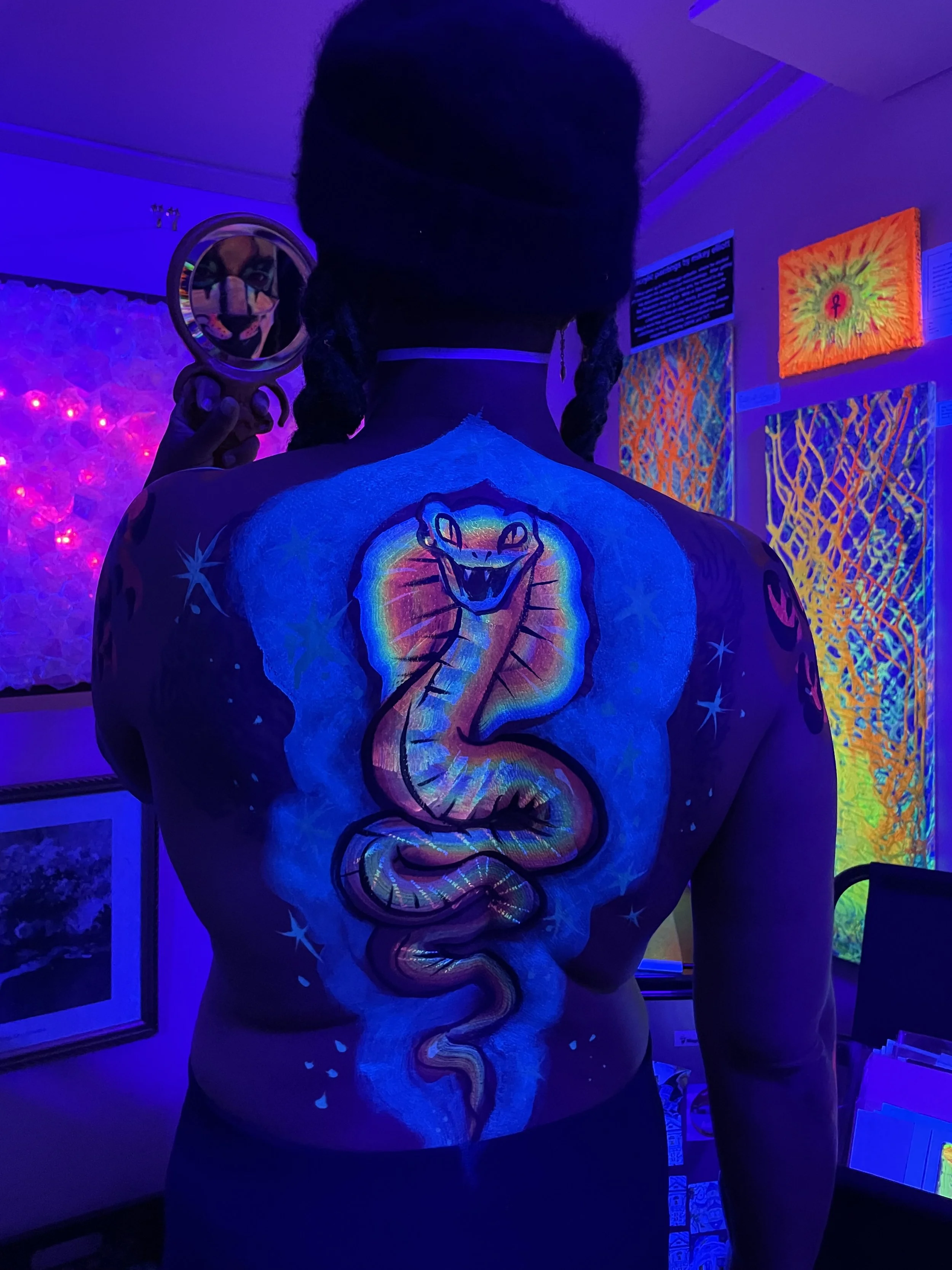 Ali Montgomery dancing before her painting “Sol’s Soul,” illuminated under black light. Her spine is painted with a glowing cobra. She holds a hand mirror that reflects her face painted with lioness features.