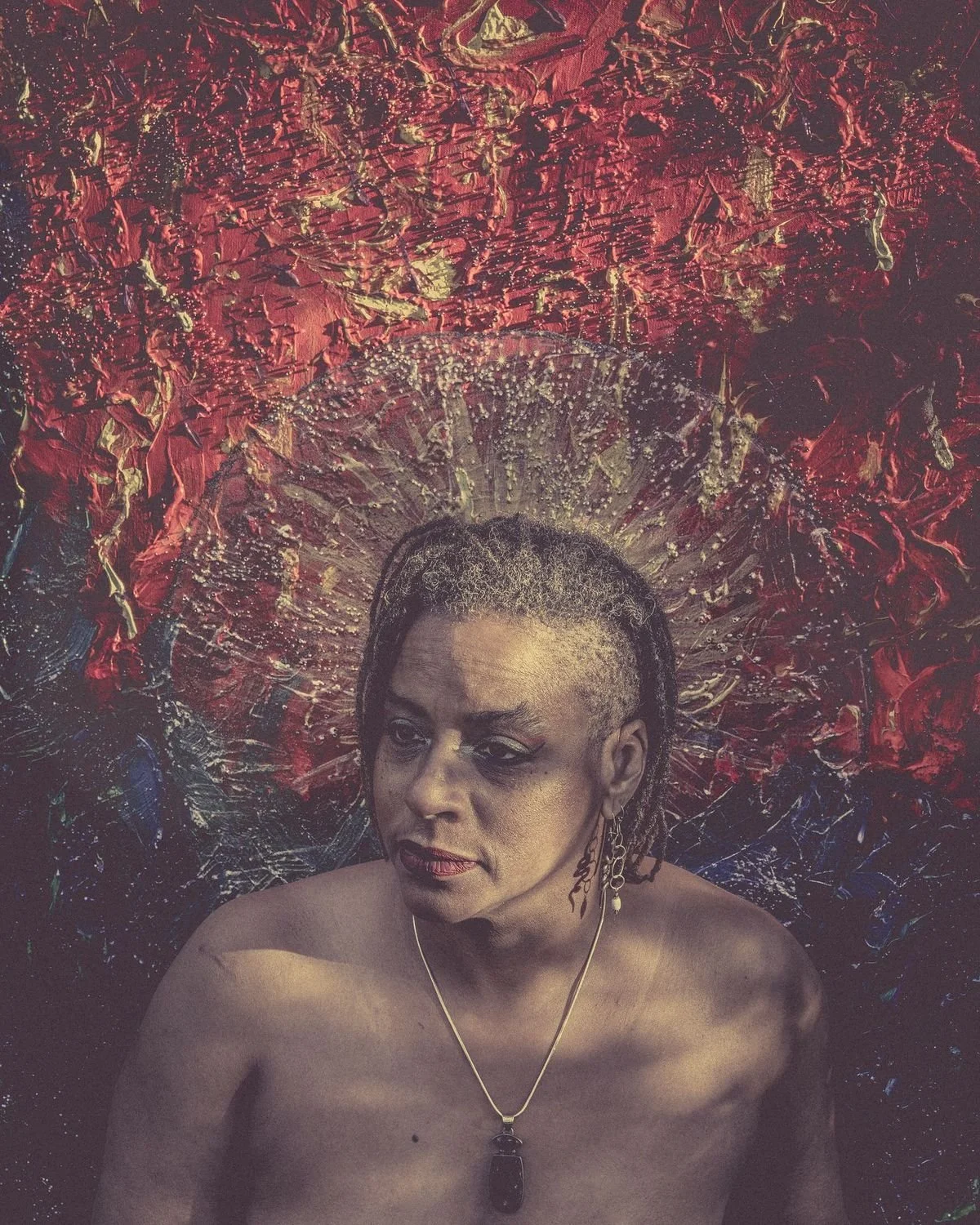 Portrait of Ali Montgomery standing before her painting Ascending the Priestess Path, with vivid red, gold, and blue behind her, posed in one of her Sekhmet priestess embodiments.