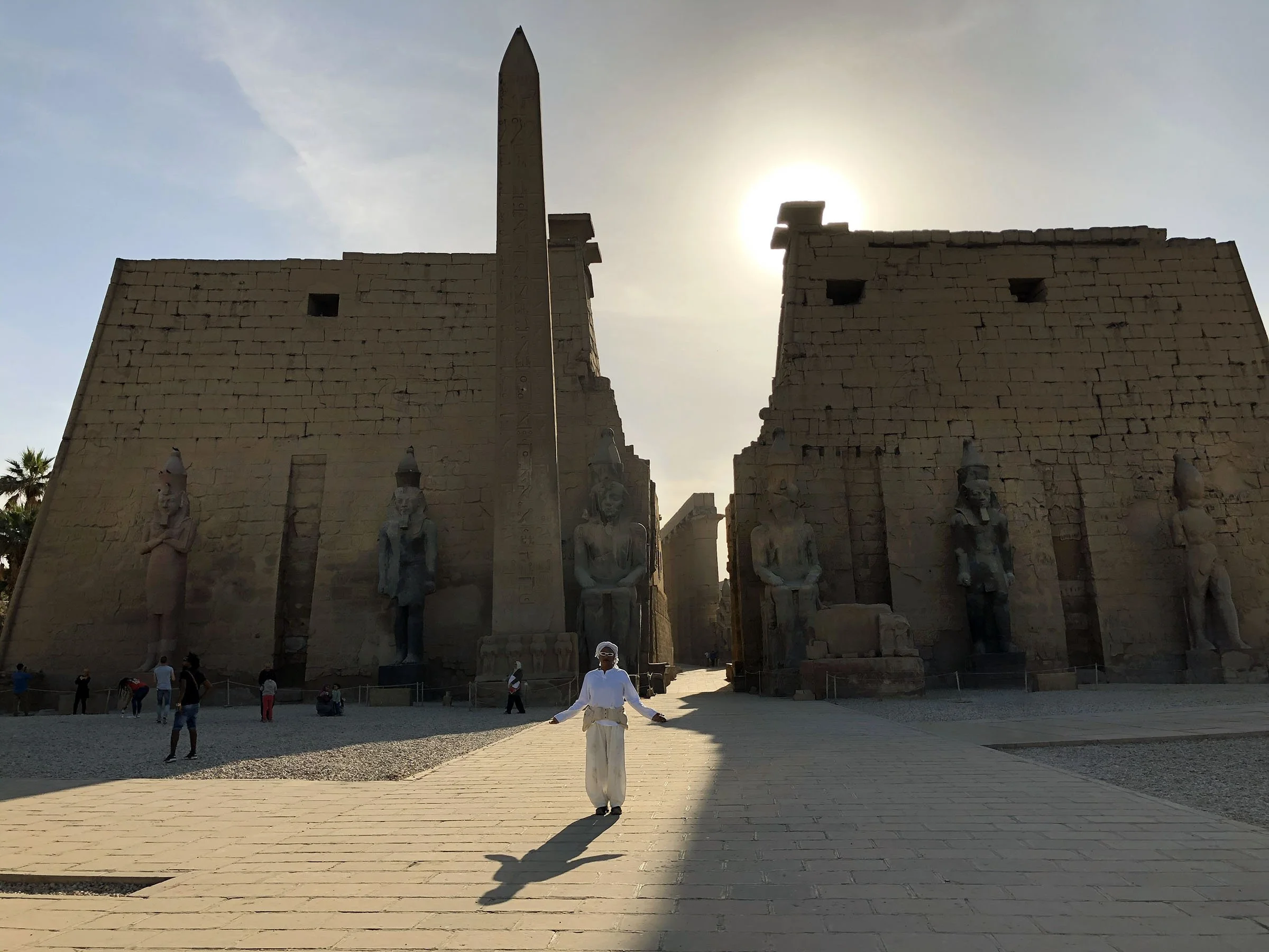 Ali Montgomery before great stone temple entrance at Luxor with six colossal pharaoh statues and a tall obelisk, backlit by the rising sun over the temple wall.