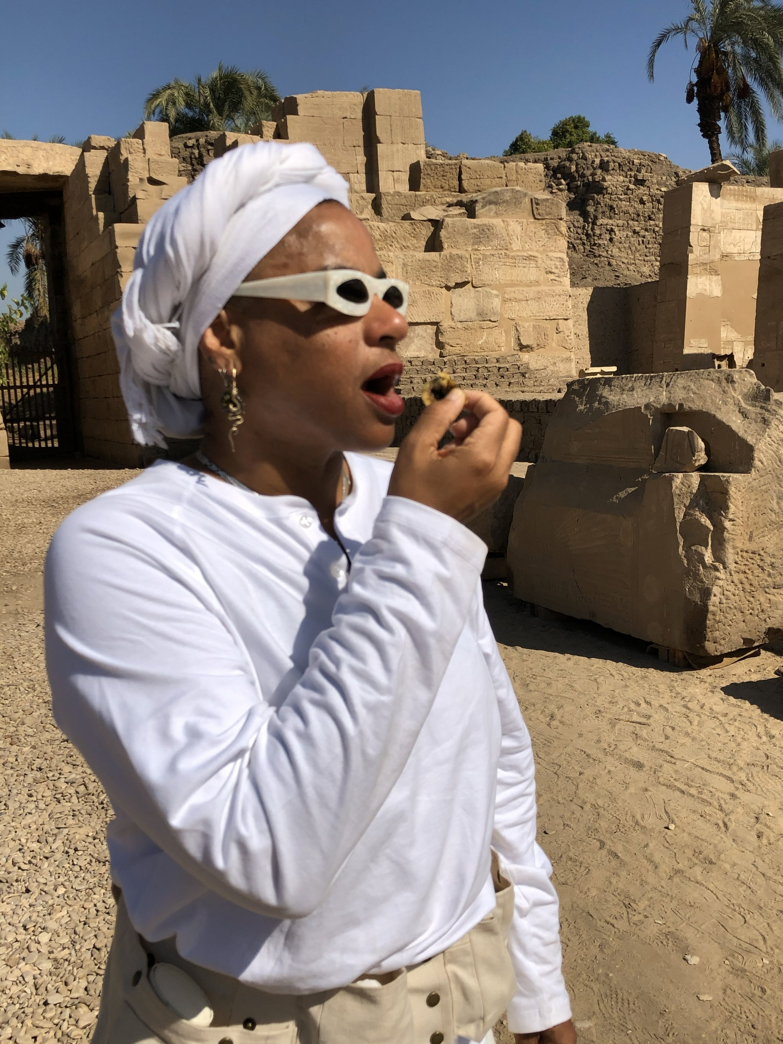 Ali Montgomery in white ritual garb and white sunglasses eating a small fruit in the courtyard outside the Temple of Ptah at Karnak.
