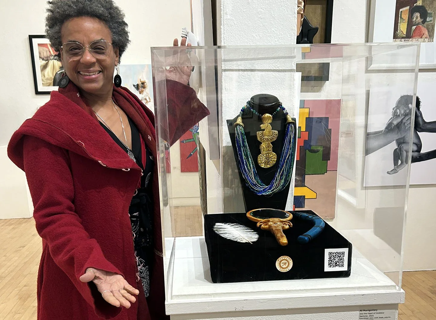 Ali Montgomery standing at the Richmond Art Center beside a plexiglass display case holding her ritual implements: a gold-colored mirror with Hathor-head design, a blue throw-stick marked with udjat eye and lotus symbols, a white ostrich feather, and