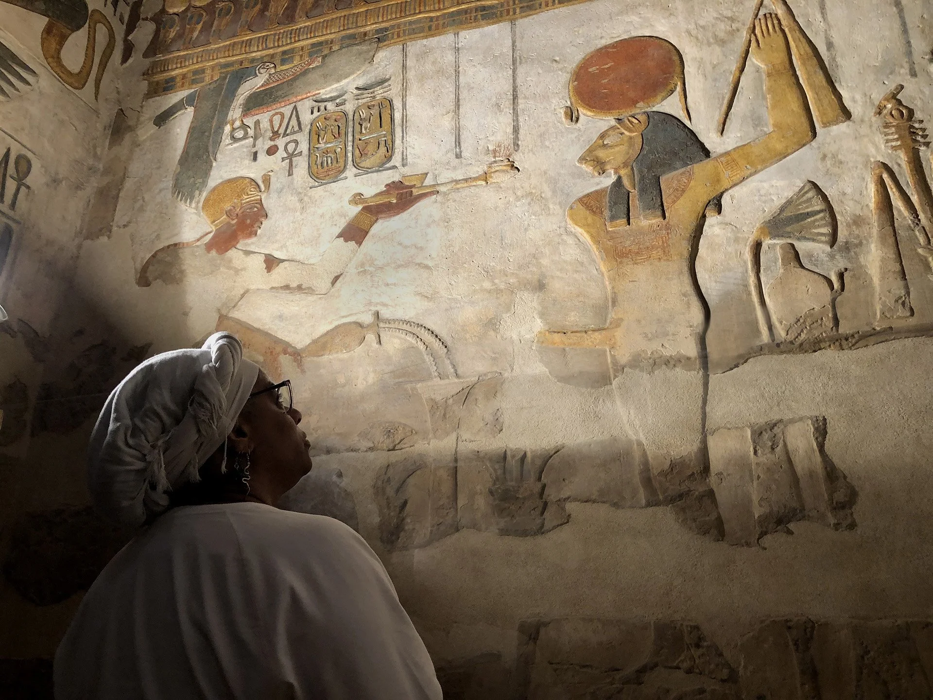 Ali Montgomery, wearing a white headwrap, stands before a painted wall relief in the Khonsu Temple at Karnak. The carving shows a lion‑headed deity receiving an offering from a pharaoh, surrounded by hieroglyphs and sacred symbols.