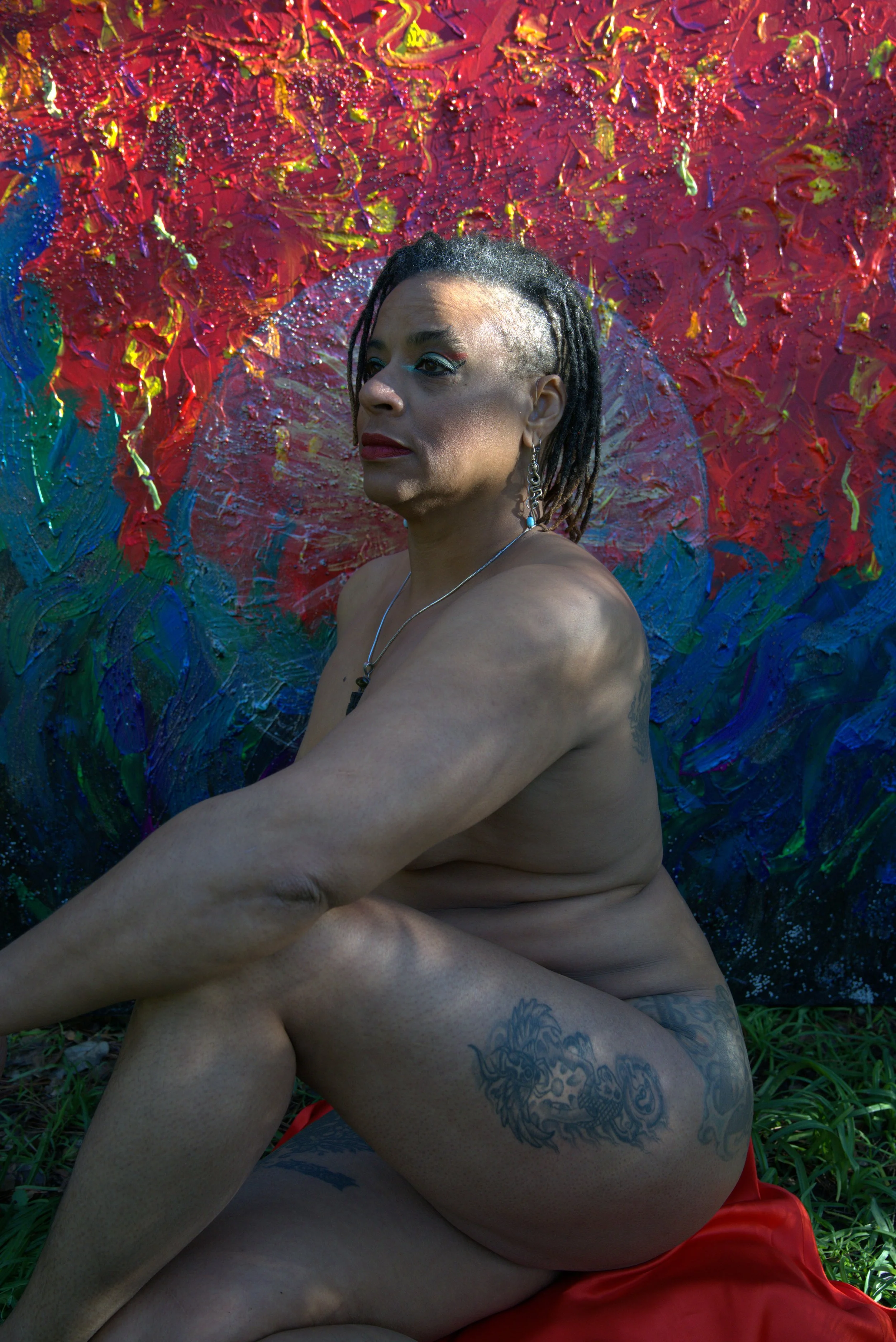 Ali Montgomery in profile sits on a red cloth outdoors, arms wrapped around knees, with visible tattoos and jewelry. Behind her is her painting “Ascending the Priestess Path,” a vibrant abstract work with bold strokes of red, blue, green, and yellow.