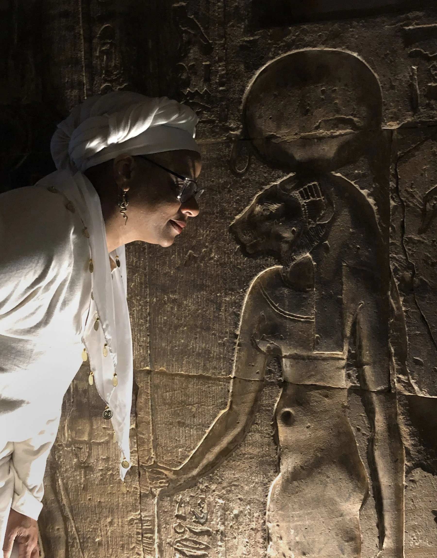 Ali Montgomery in white ritual garb standing face‑to‑face with a wall relief of the Goddess Sekhmet in a dark interior corridor of the Temple of Horus.
