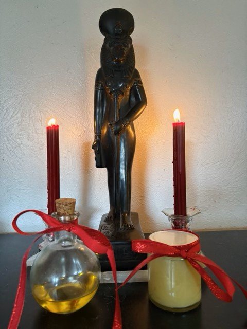 Home altar with a Sekhmet statue, two red candles, and bottles of oil and unguent tied with red ribbons.