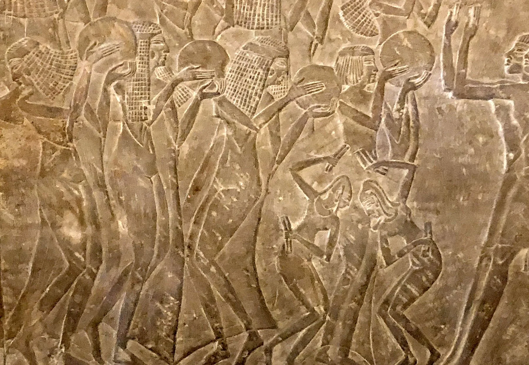Flat stone bas‑relief showing a procession of women in profile, dancing and holding round hand cymbals. The figures wear stylized garments and hairstyles, suggesting a ceremonial scene.
