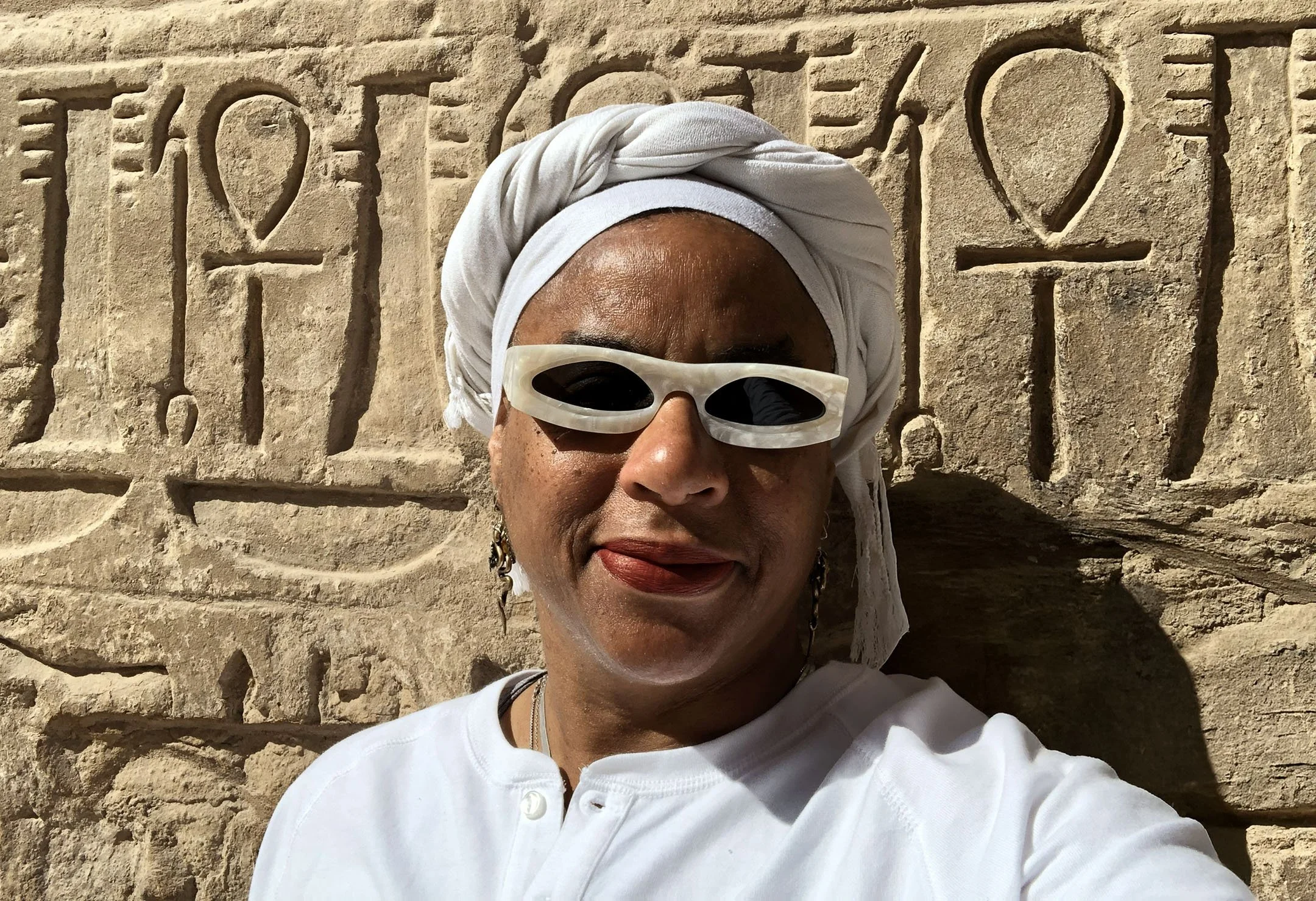 Ali Montgomery in white ritual garb and sunglasses standing before a sunlit temple wall carved with multiple ankhs and hieroglyphs.