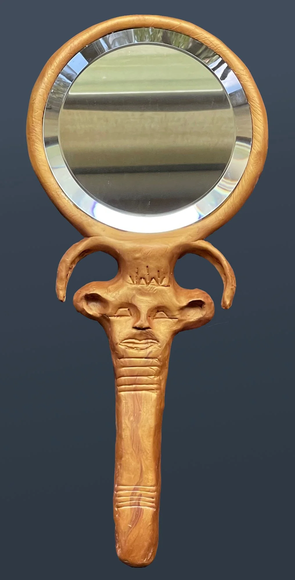Hand mirror with circular reflective surface. The gold-colored handle is sculpted in a Hathor-head design, showing a stylized face with prominent ears and curved horn-like extensions above the head.