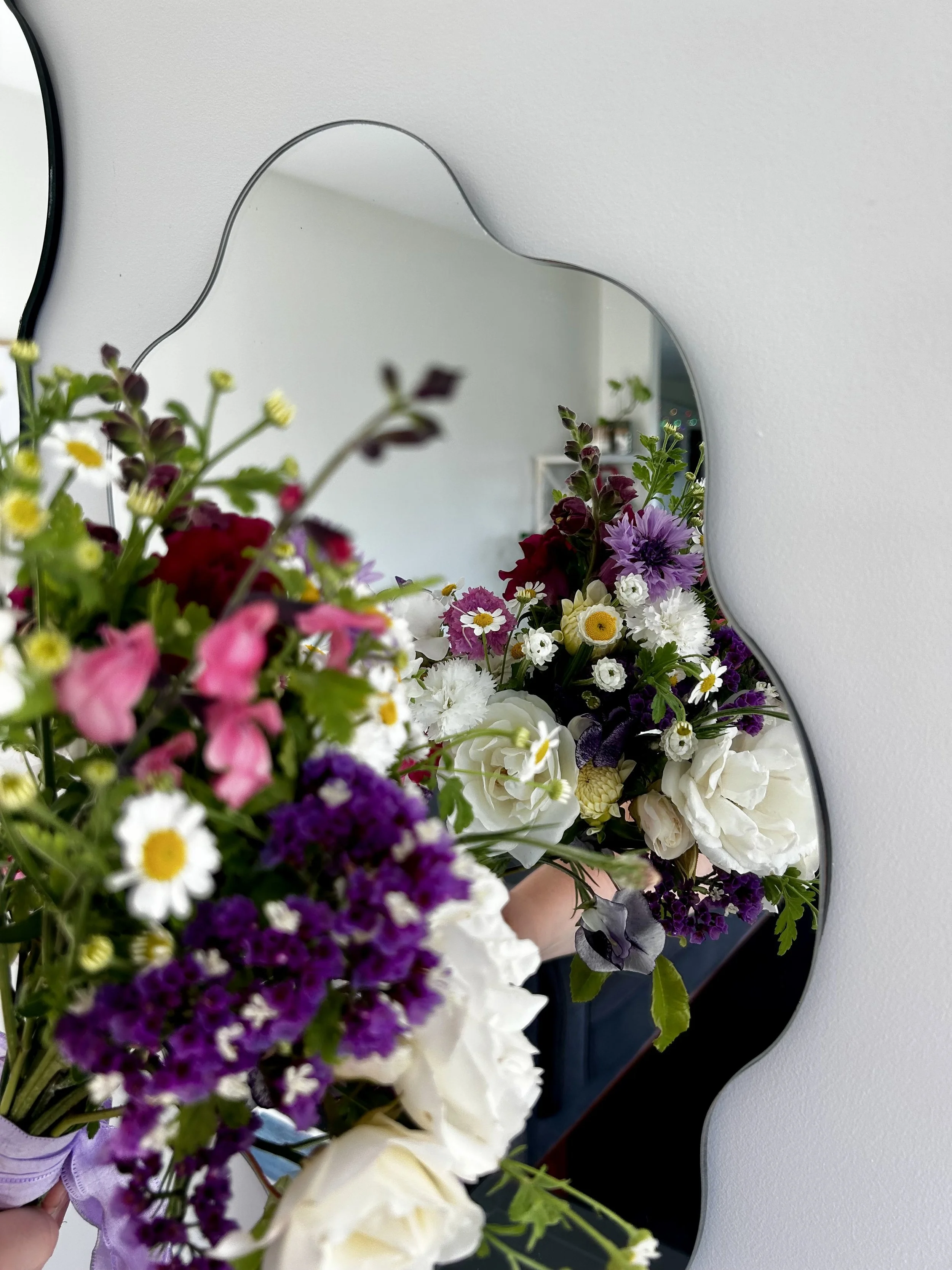 A mirror on a white wall reflects a person holding a colorful bouquet of flowers with various types and colors including white, purple, yellow, and pink.