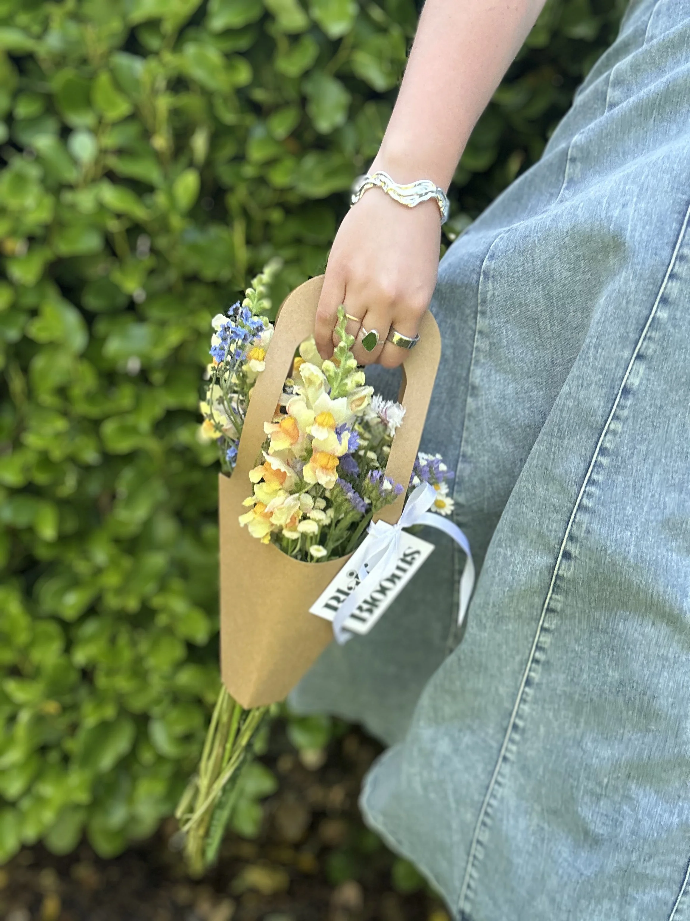 Person holding a small bouquet of colorful flowers in a paper wrapper outdoors, wearing a silver bracelet and rings.