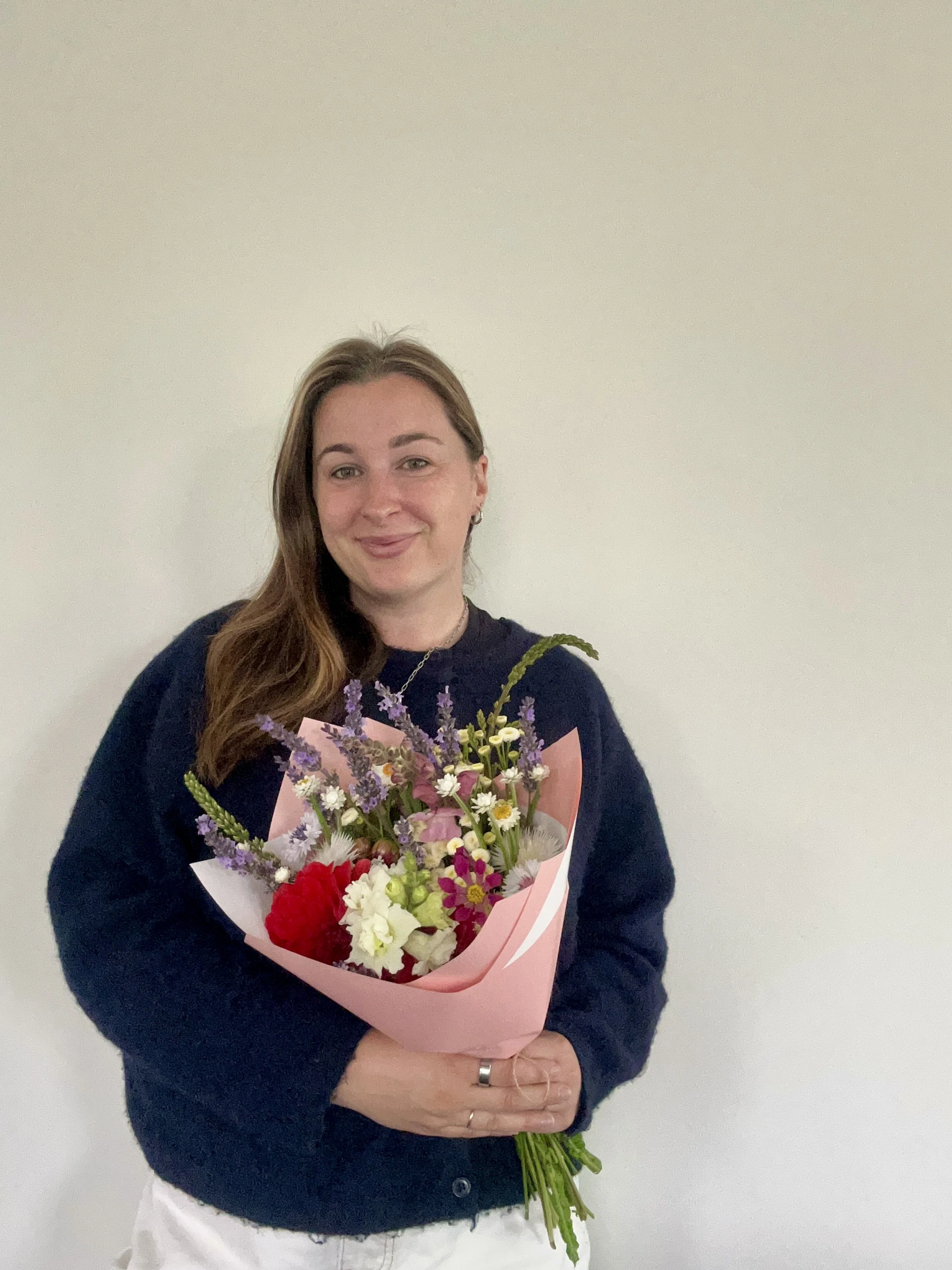 A woman with long brown hair holding a colorful bouquet of flowers, standing against a plain off-white wall.