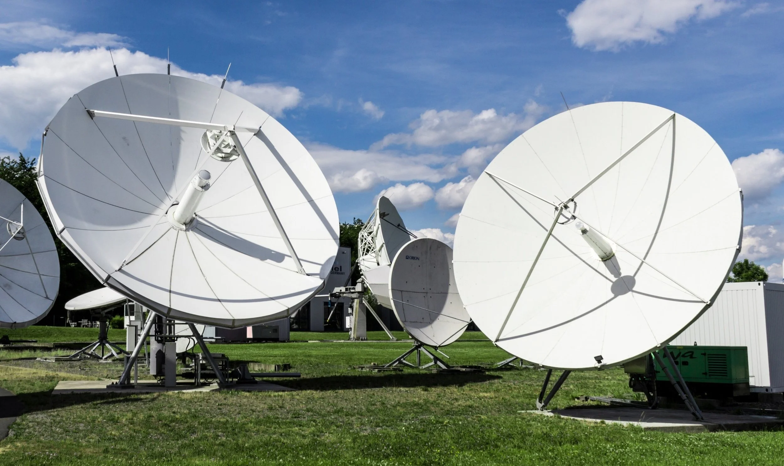 Multiple satellite dishes pointed towards the sky on green grassy ground with blue sky and scattered clouds in the background.