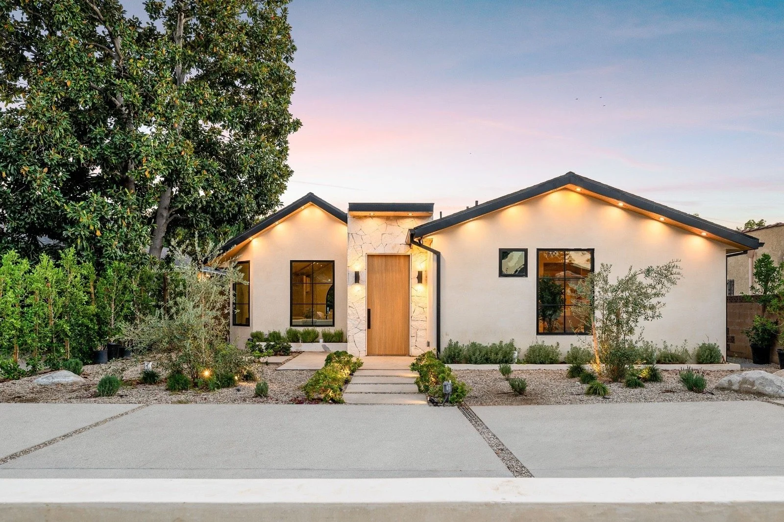 Modern single-story house with a minimalist design, stone accents, and outdoor lighting, surrounded by desert-style landscaping and a large tree on the left