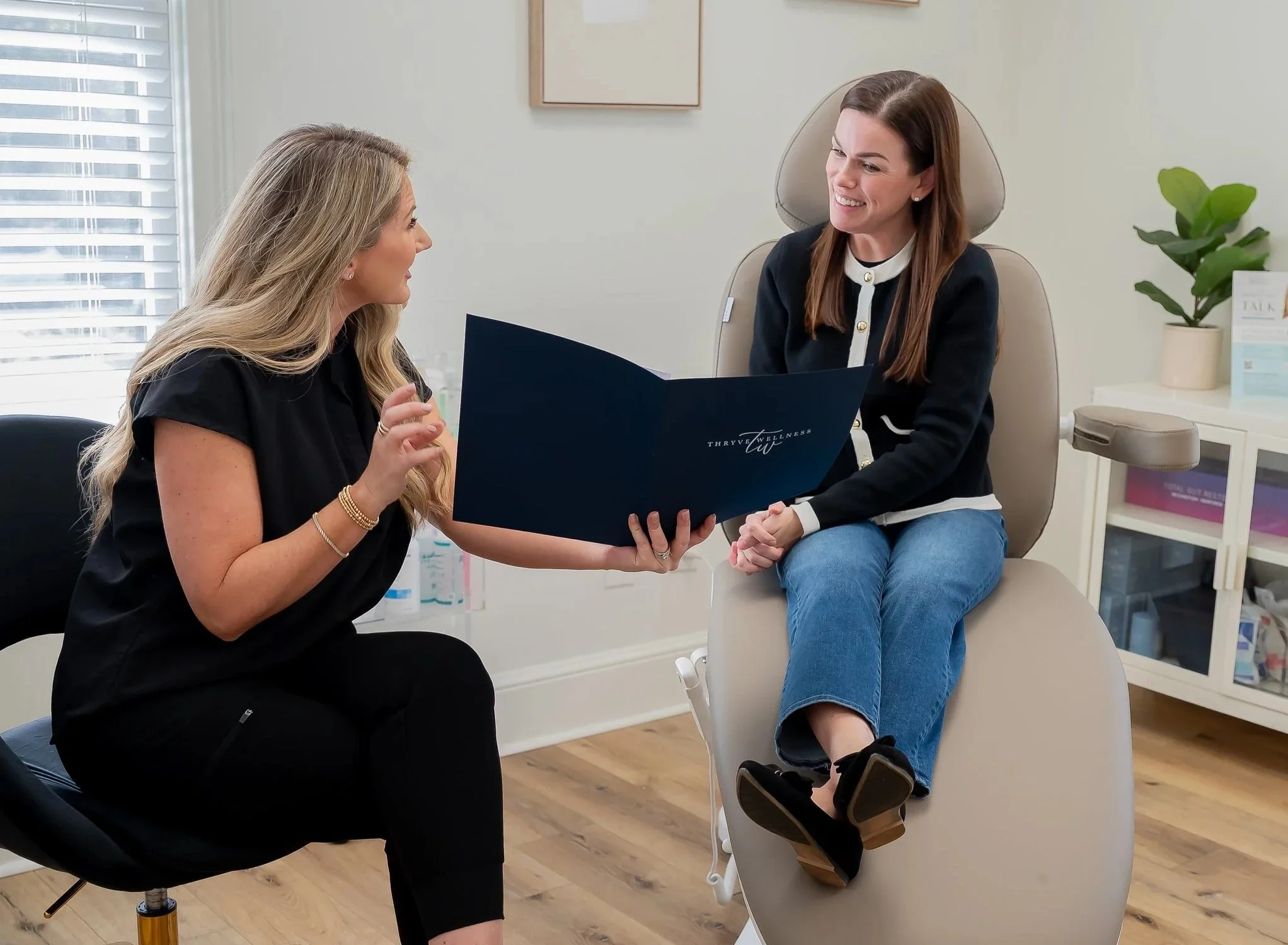 A woman sitting in a medical examination chair engaged in a discussion with a healthcare professional holding a large folder or booklet.