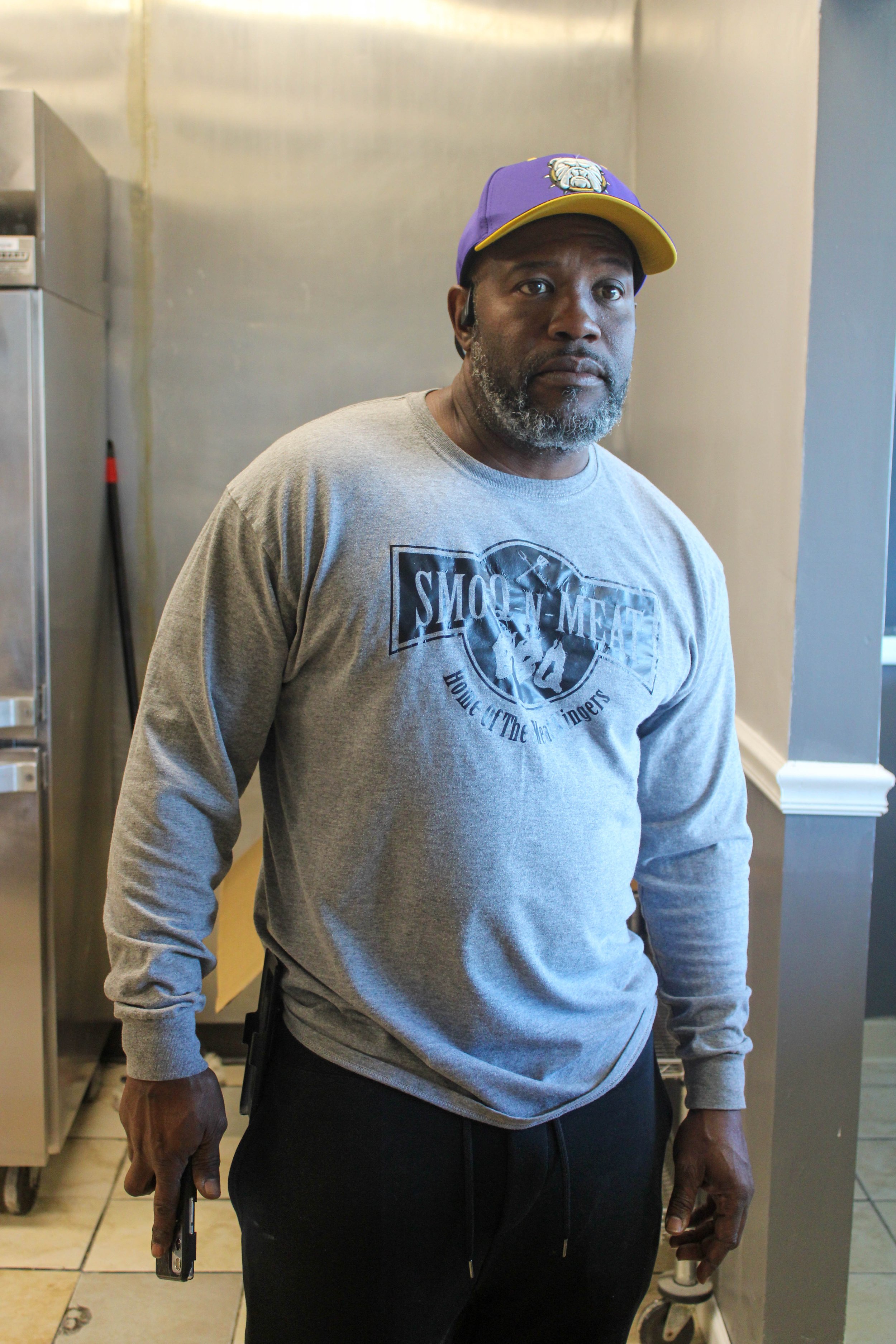 A man standing in a room, holding a phone in his right hand, wearing a purple and yellow cap, a gray long-sleeve shirt with a graphic logo, and black pants.