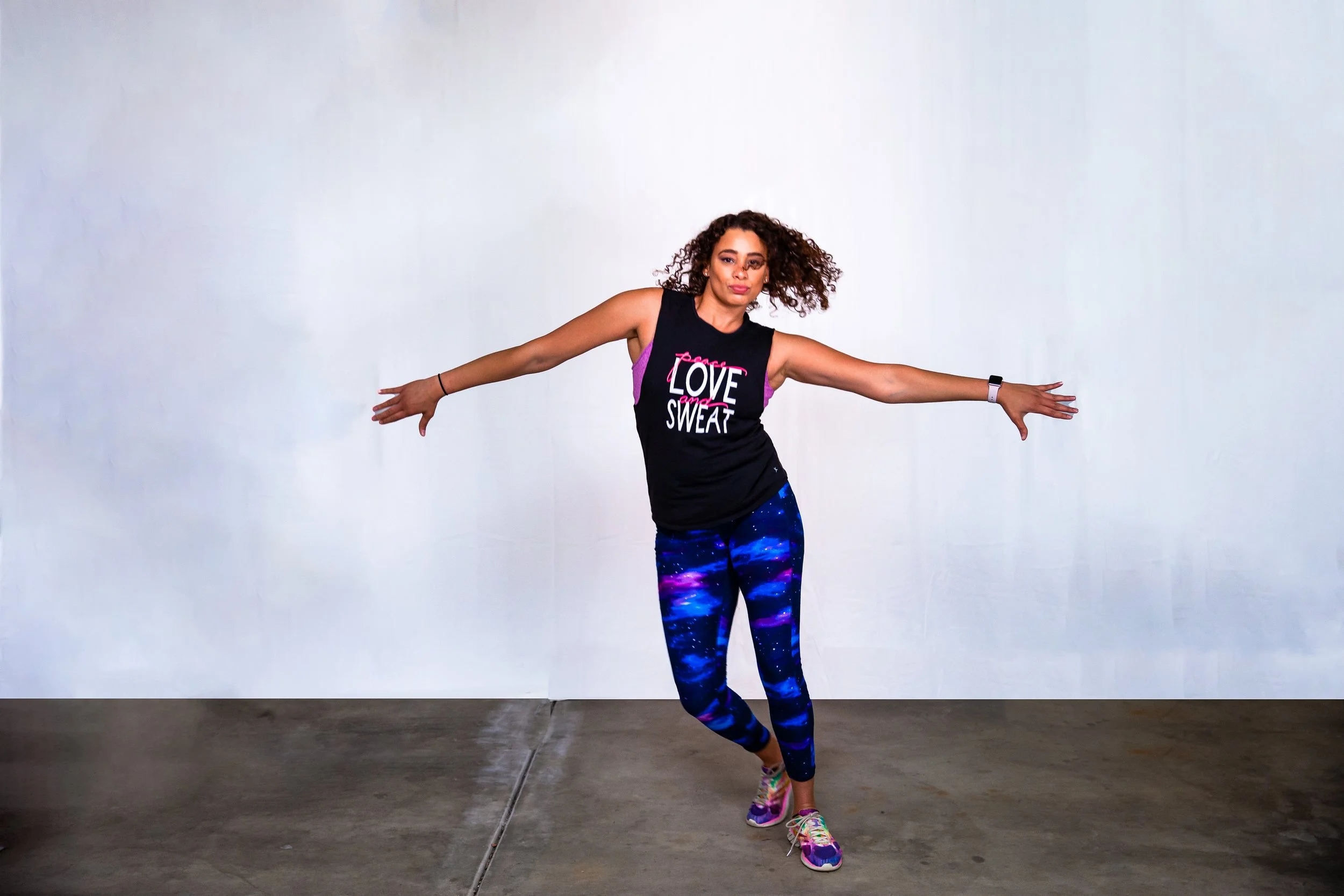 A woman with curly hair dancing or stretching in front of a plain white wall, wearing a black tank top with the words "Love and Sweat" and colorful galaxy-patterned leggings.