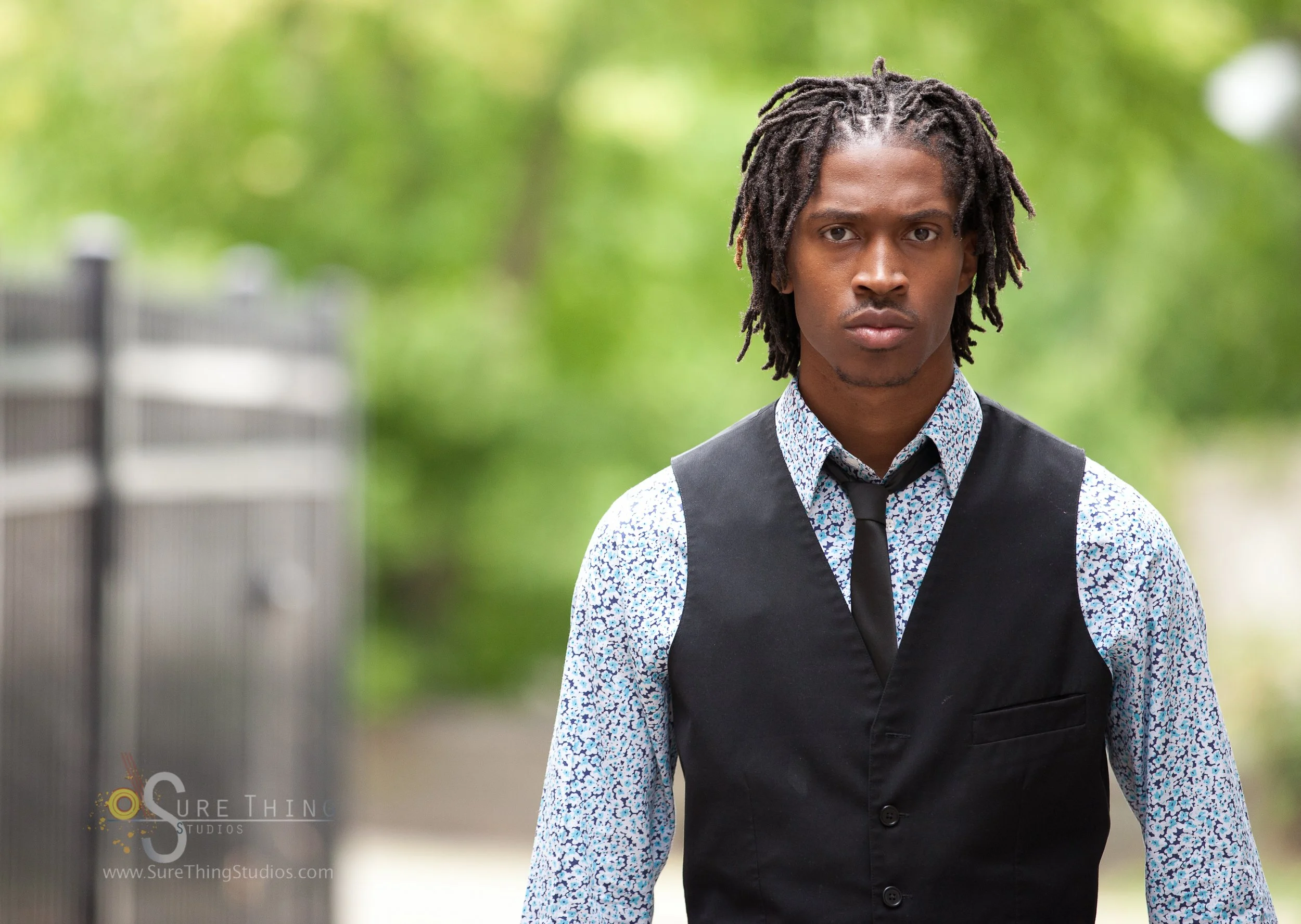 A young man with dreadlocks wearing a patterned shirt, black vest, and tie standing outdoors with green foliage in the background