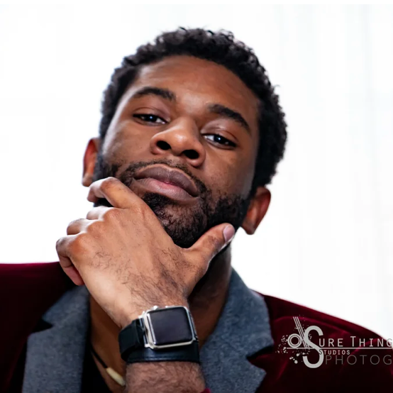 A man with dark curly hair and a beard posing with his hand on his chin, wearing a dark blazer and a smartwatch.