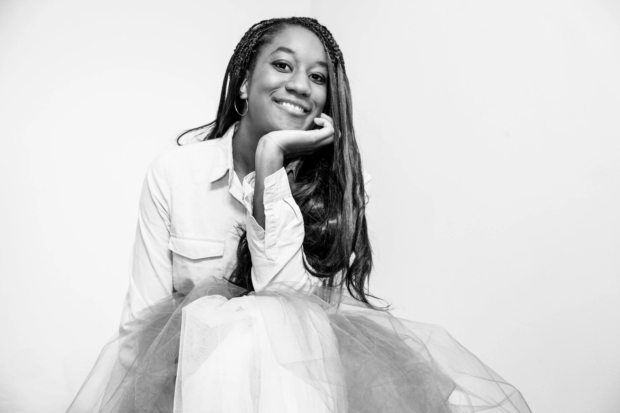 Black and white photo of a woman with long, braided hair, smiling, resting her chin on her hand, wearing a light-colored jacket and a tulle skirt.