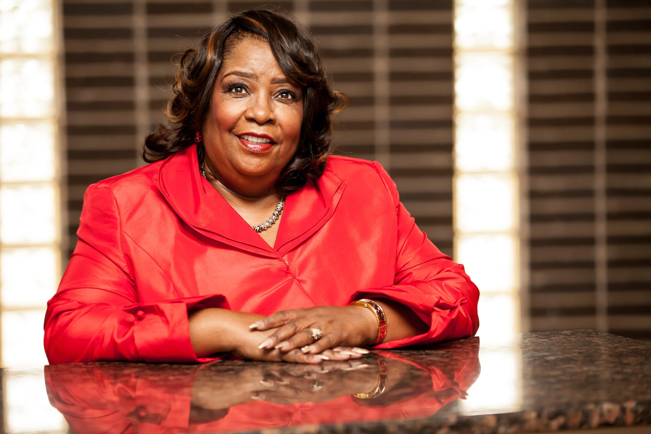 A middle-aged woman with medium skin tone, curly dark hair with highlights, smiling and wearing a red blazer, a necklace, bracelet, and ring, sitting at a polished granite table with a background of wooden blinds and light from windows.