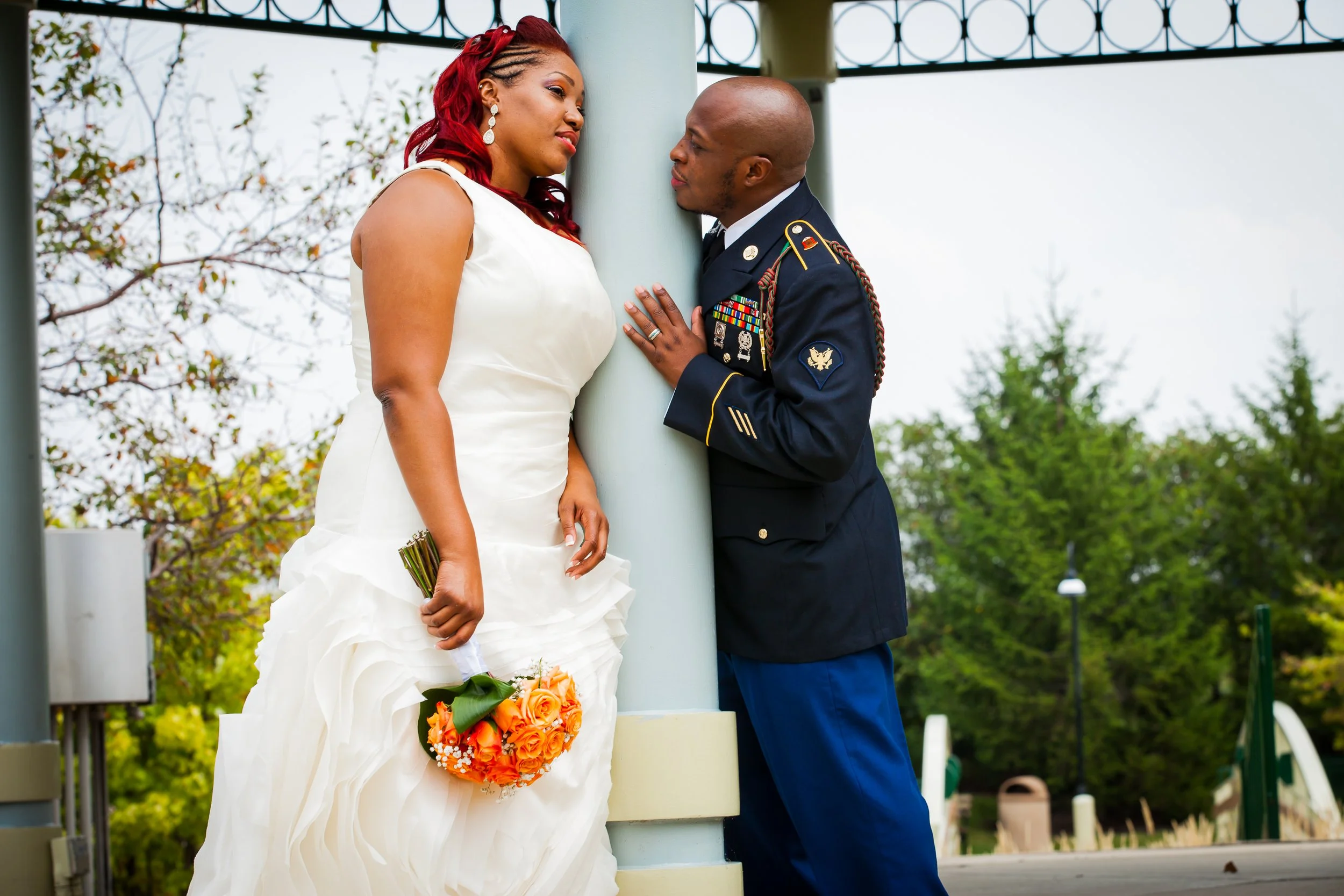 A bride in a white wedding dress holding a bouquet of orange roses standing close to a man in a military uniform, with both leaning against a light blue support column outdoors with trees in the background.