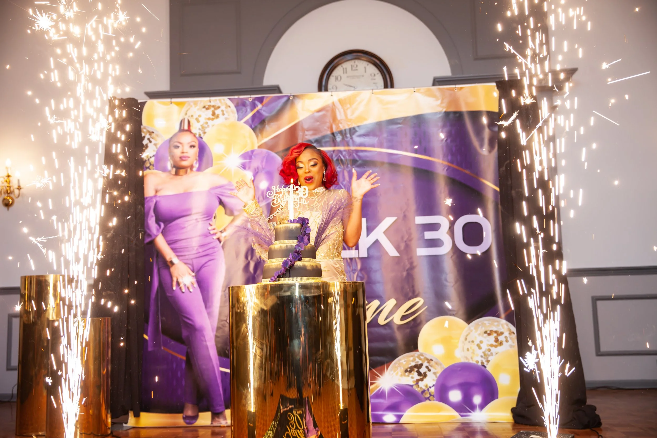 A woman with red hair celebrating her 30th birthday with a cake, sparklers, and balloons in the background.