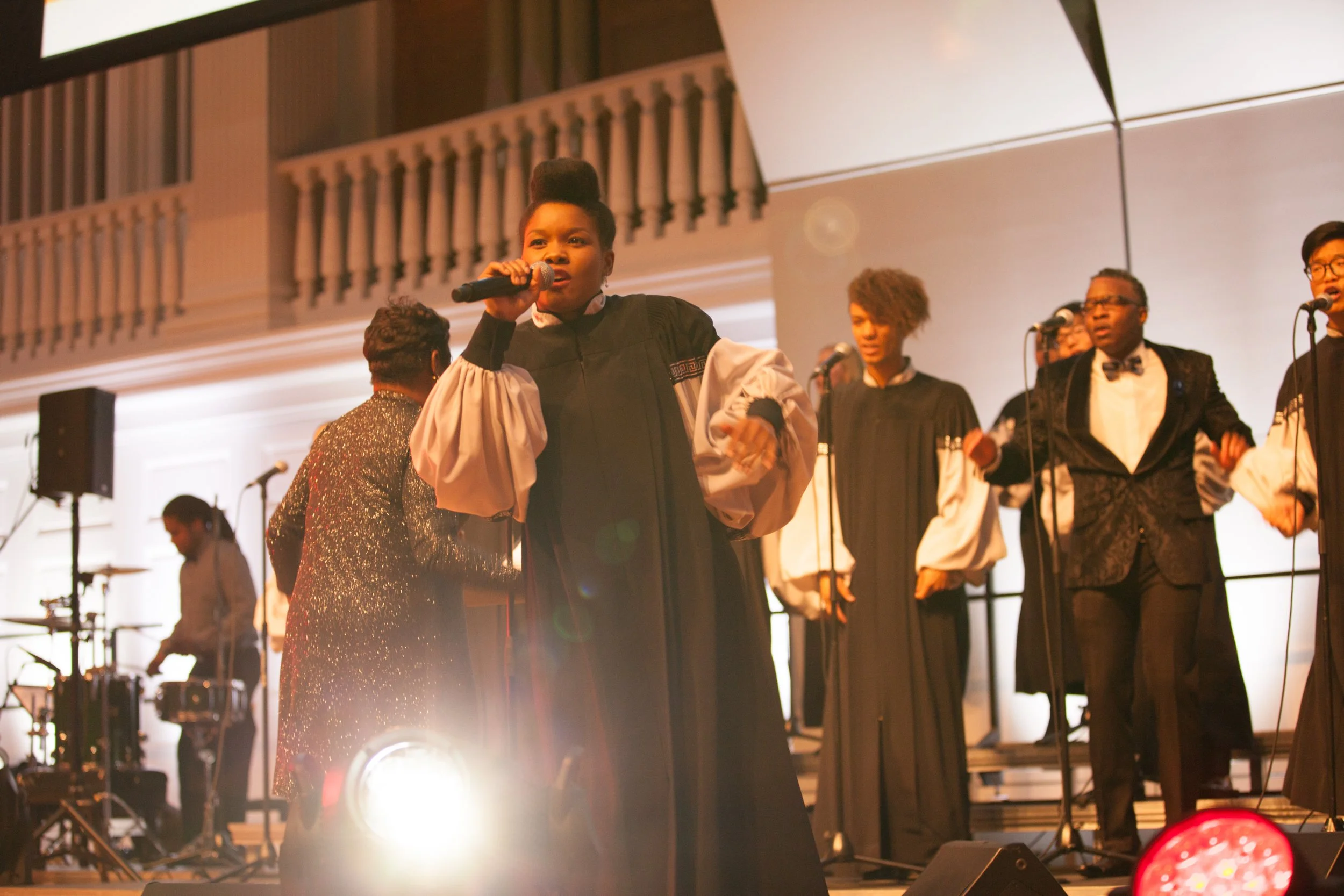 A group of singers and musicians performing on stage at a formal event. The lead singer, a woman wearing a black and white choir robe, is holding a microphone and singing. Behind her are backup singers, some in similar choir robes and one in a tuxedo. In the background, a person is playing drums, and stage lighting illuminates the scene.