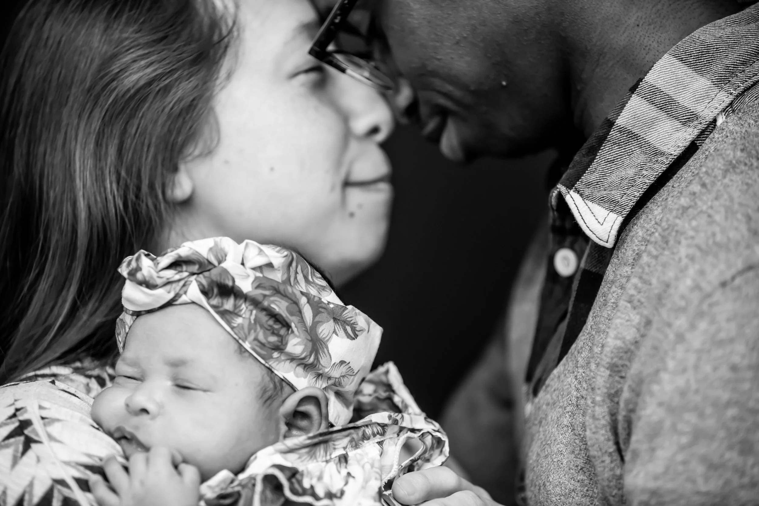 Black and white photo of a family showing a woman and a man leaning their foreheads together with their eyes closed, and a sleeping baby resting on the woman's shoulder.
