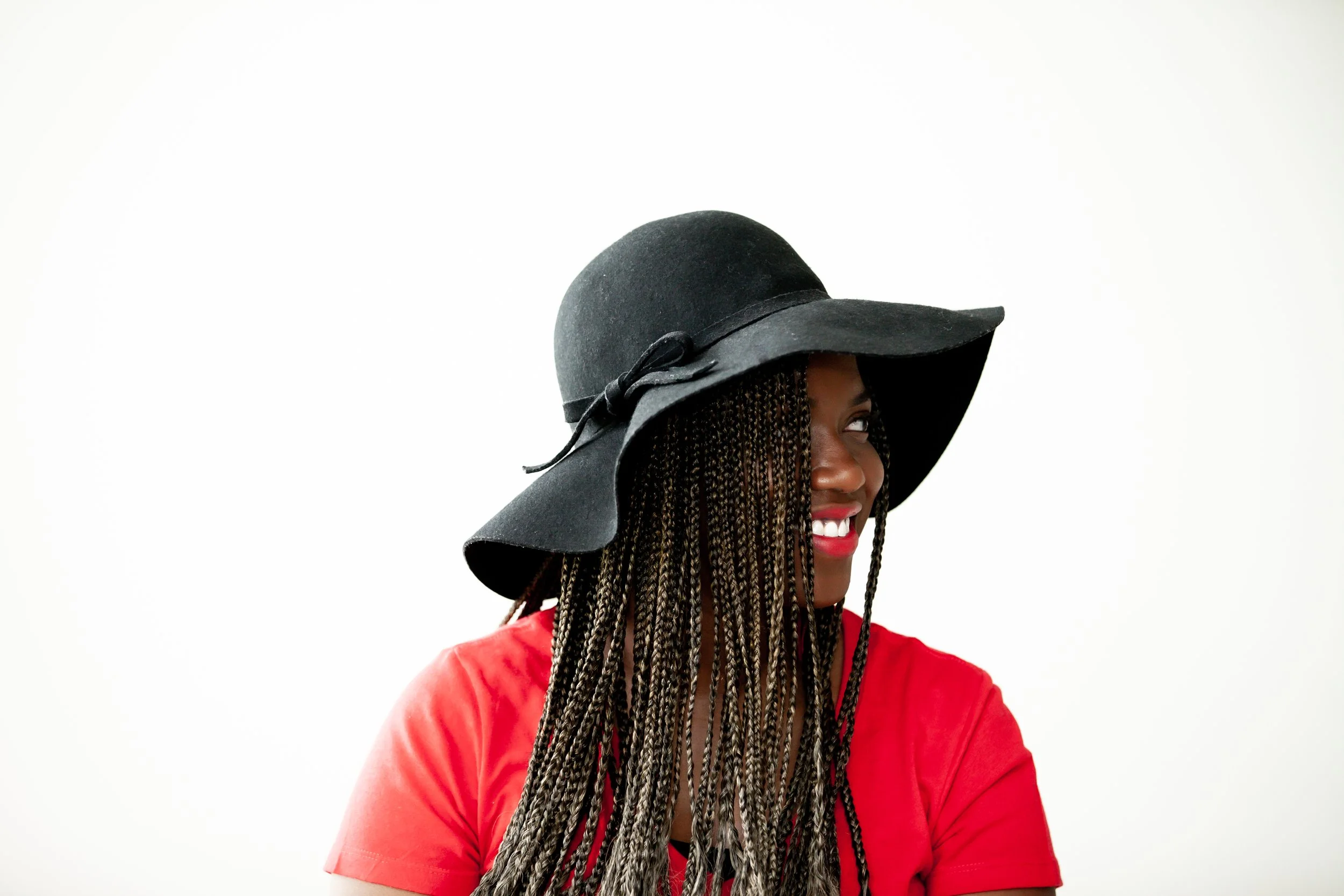 A woman in a black floppy hat with a decorative ribbon, a red shirt, and long braided hair, smiling and looking to her right against a plain white background.