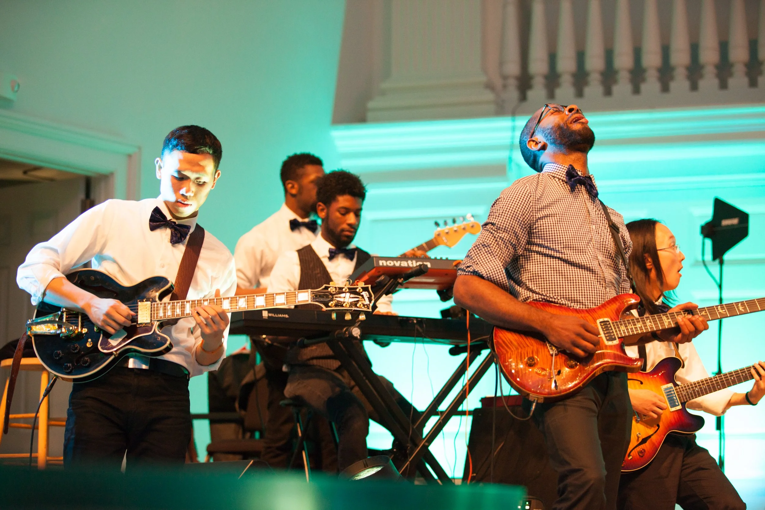 Group of musicians performing on stage, playing guitars and keyboard, in a formal setting with colorful lighting.