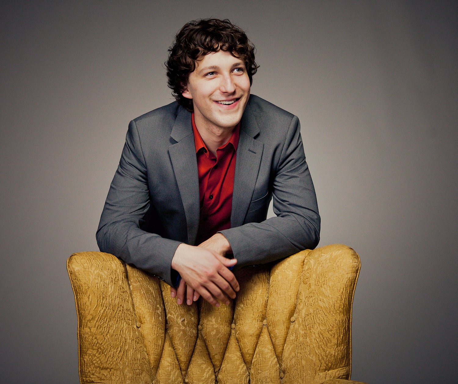 A young man in a gray suit with a red shirt, smiling and leaning on a gold velvet chair with a tufted back.