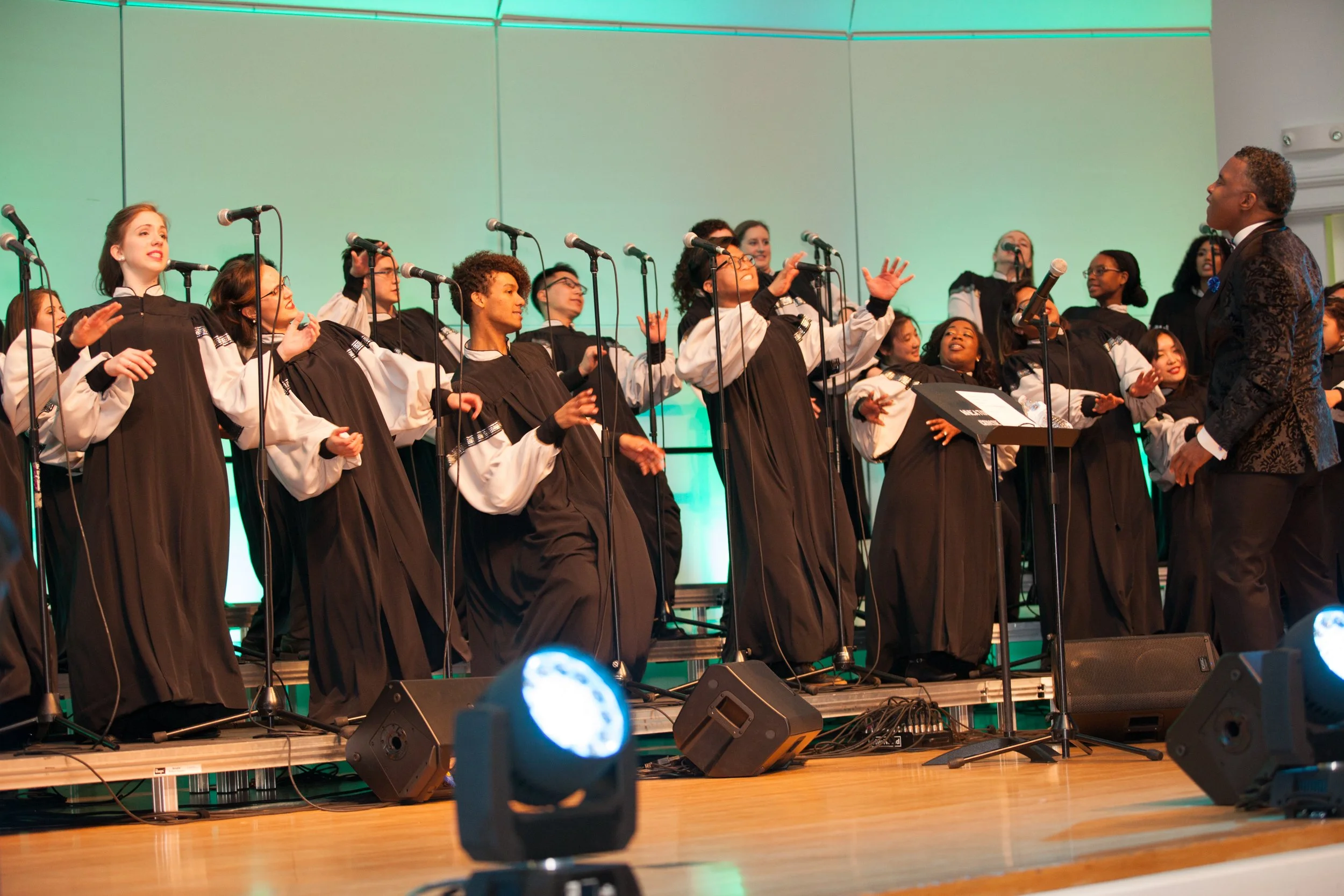 Choir of young singers dressed in black robes with white sleeves, performing on stage with a conductor, microphones, and stage lights.