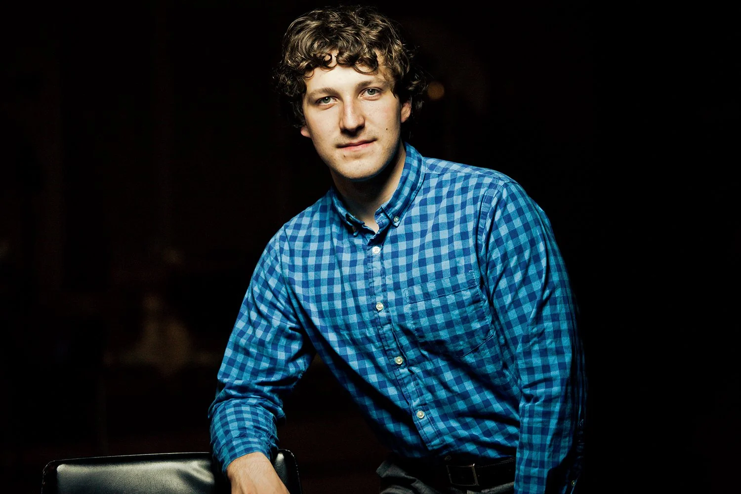 Young man with curly hair in a blue checkered shirt, sitting against a dark background.