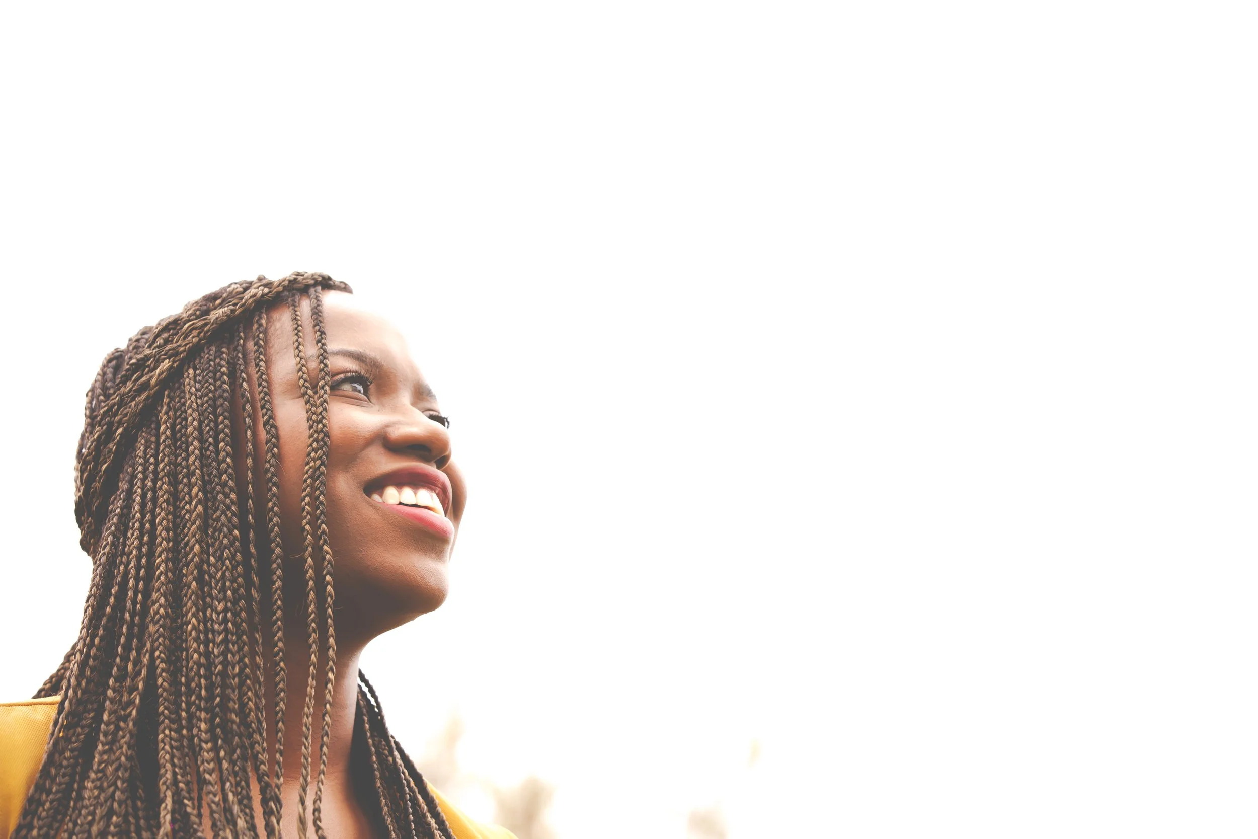 A young Black woman with long braided hair smiling and looking up against a bright white sky.