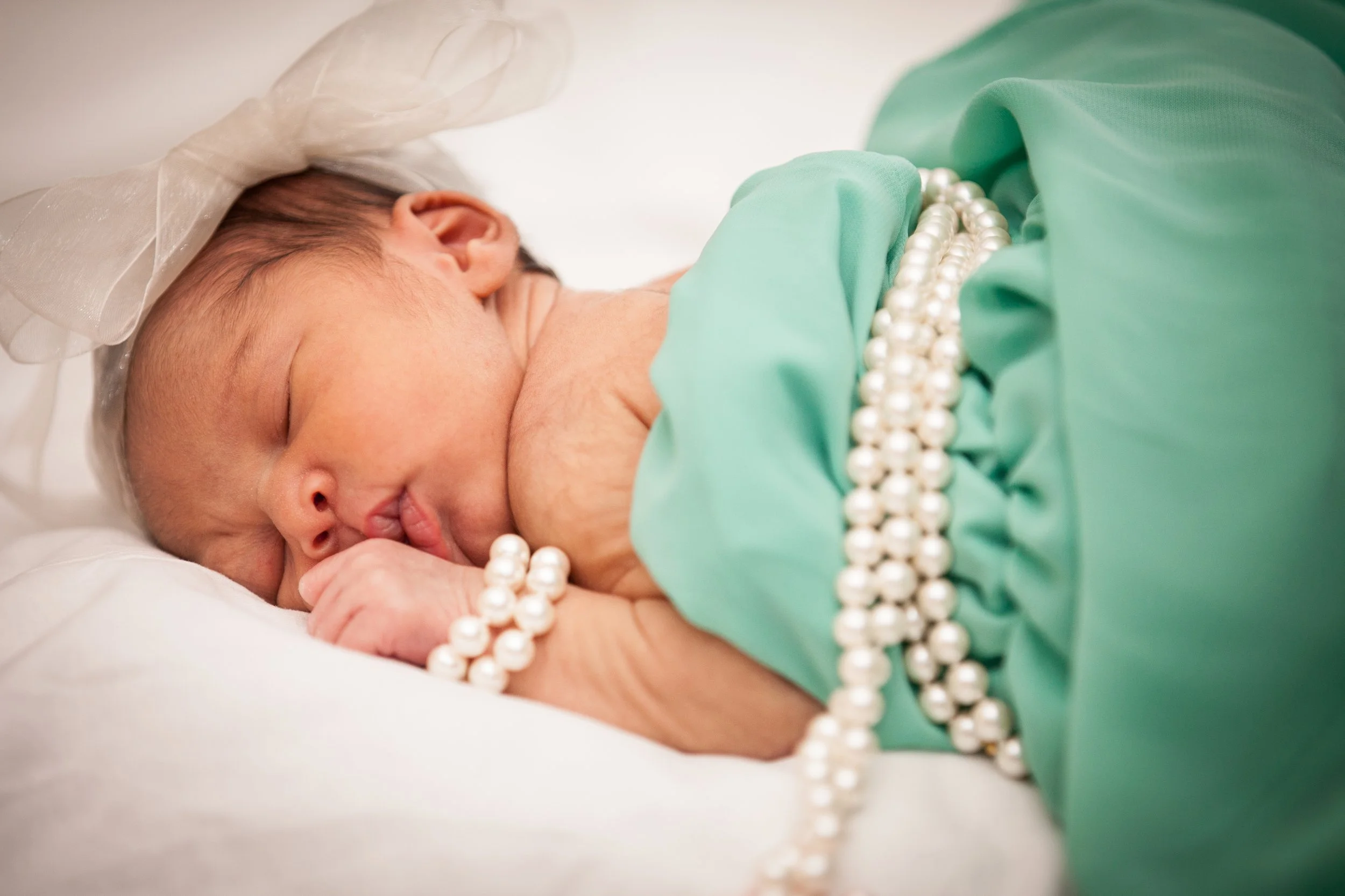 A sleeping baby with a pearl bracelet, wearing a green dress and a headband, resting on a white surface.