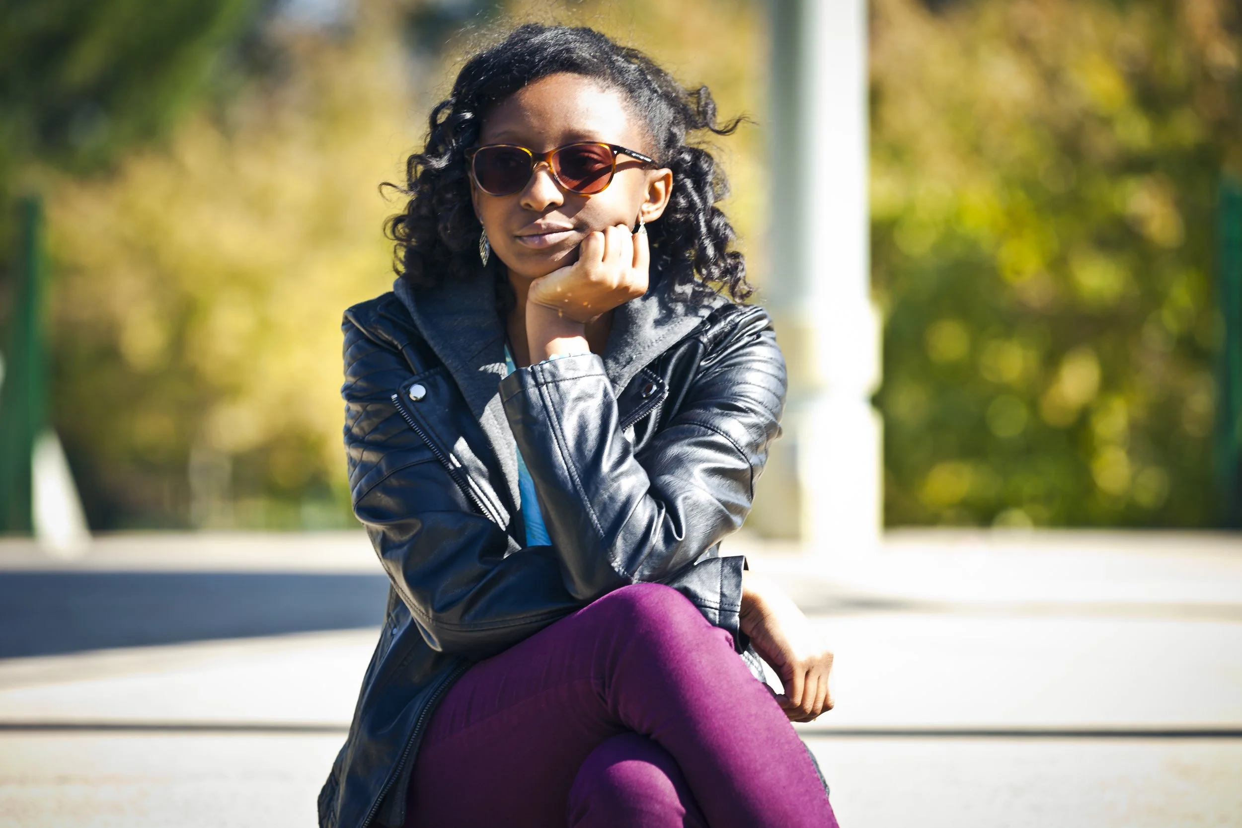 A woman wearing sunglasses, a black leather jacket, gray hoodie, and purple pants sitting outdoors on a bright day with trees in the background.