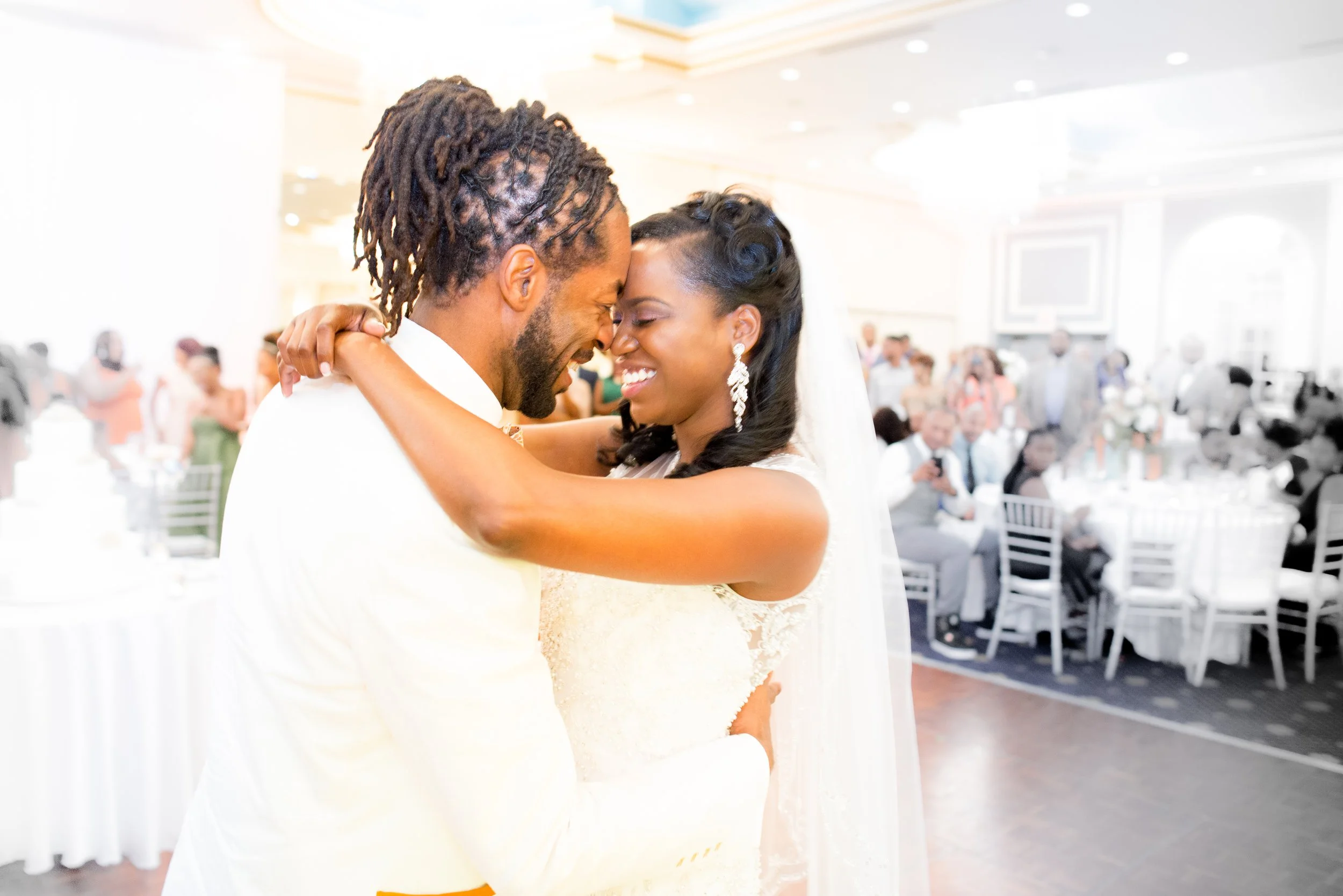 A bride and groom share a dance at their wedding reception, smiling with their foreheads touching, in a bright, elegant venue filled with seated guests.