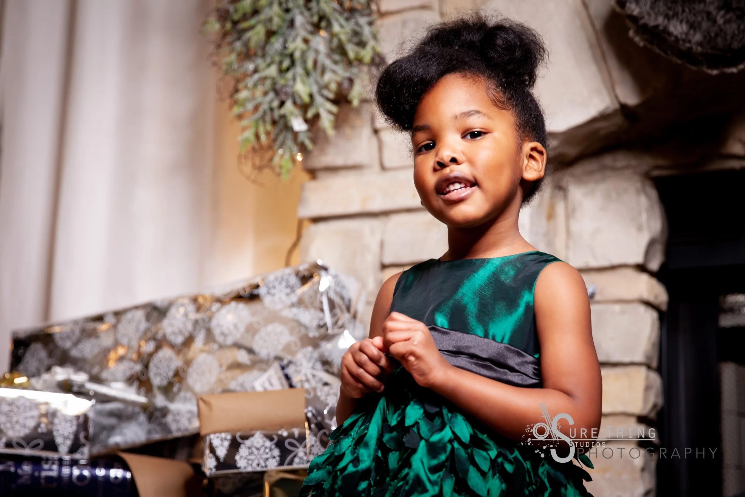 A young girl with dark curly hair styled in an updo, wearing a green and black sleeveless dress, standing in front of a stone fireplace with wrapped Christmas presents nearby.