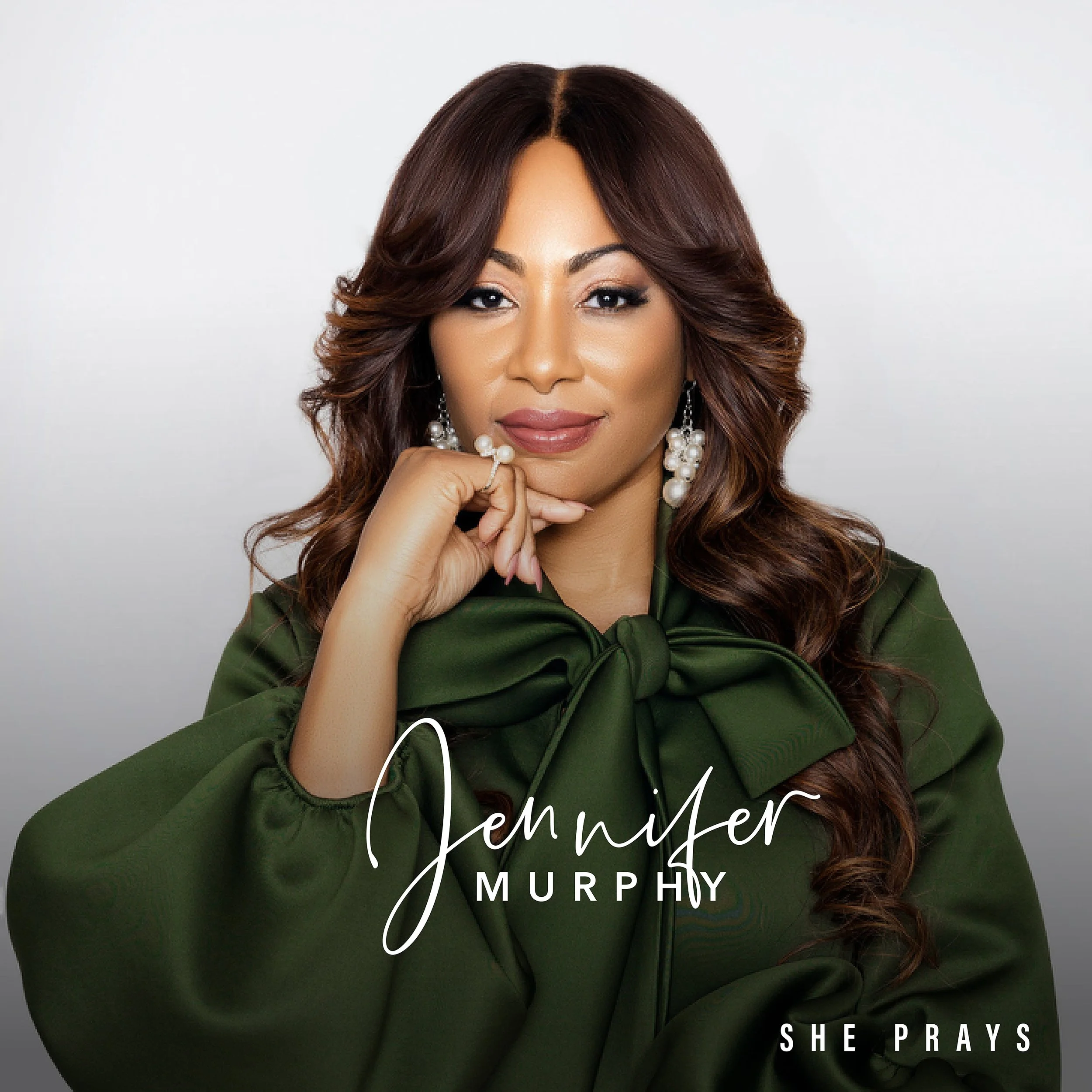 A woman with long wavy brown hair wearing a green blouse with a large bow and pearl earrings, resting her chin on her hand, on a light background. Text on the image reads 'Jemimah Murphy' and 'She Prays'.