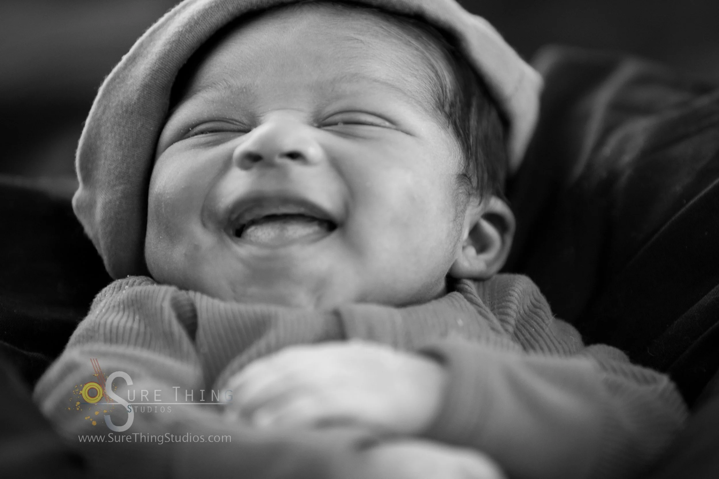 Black and white photo of a smiling baby wearing a hat and a cozy outfit, lying down.